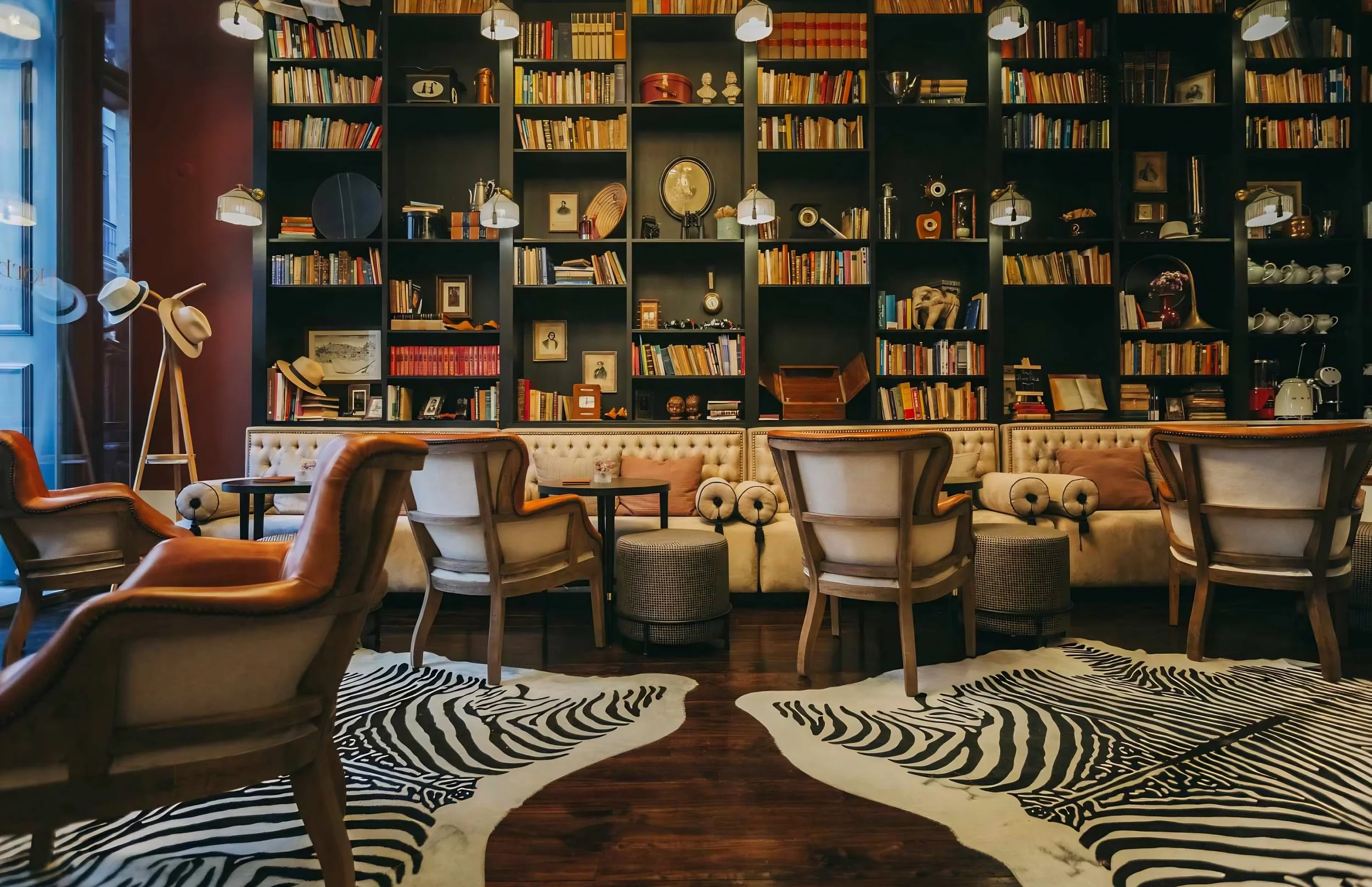 Interior of a cozy library or reading room with a large black bookshelf filled with books and decorative items. There are multiple plush armchairs arranged around a zebra-print rug, and a beige tufted sofa with pillows. A floor lamp with hats and a zebra-patterned rug are also visible.