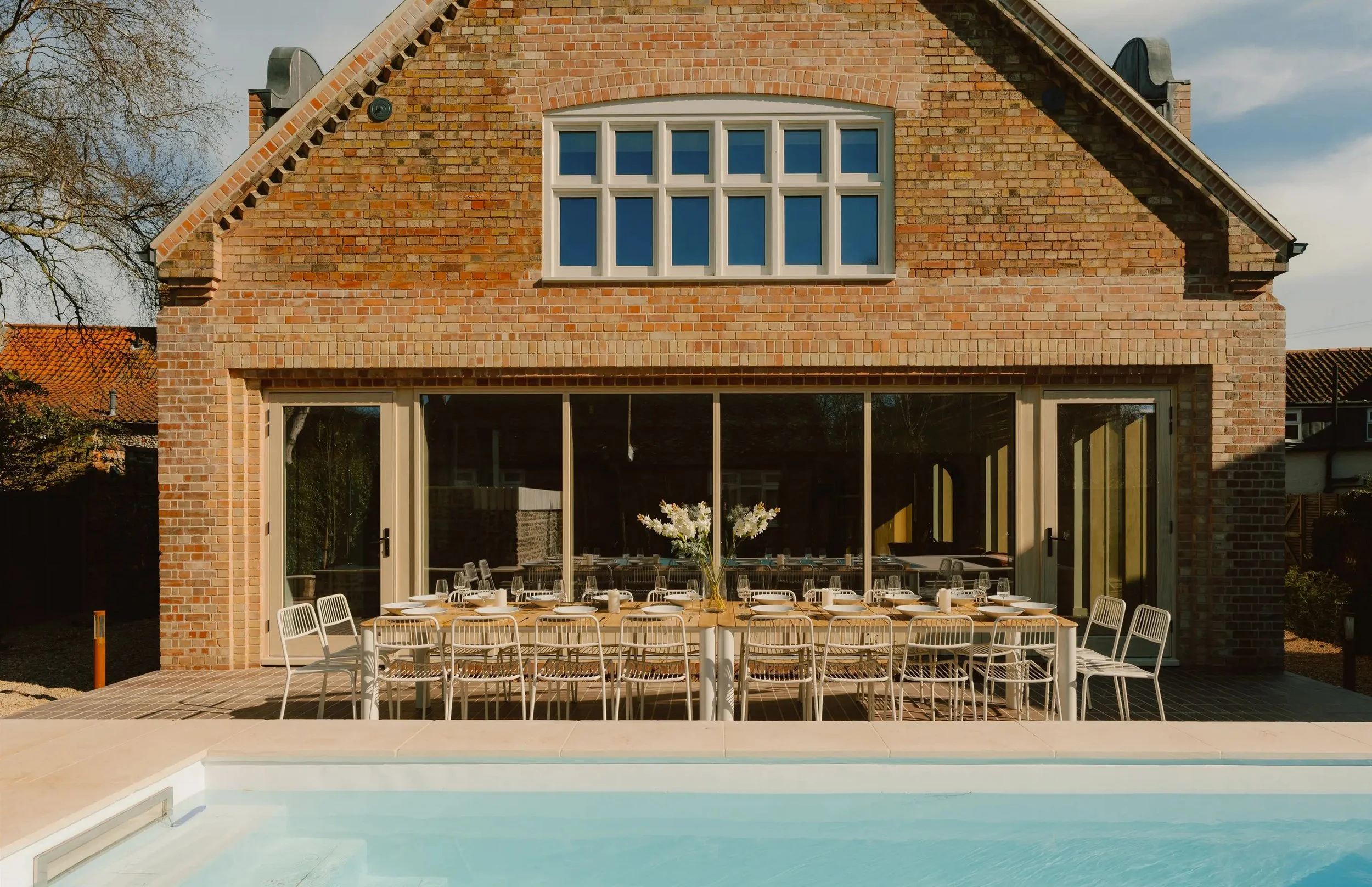 Backyard patio with dining table set for a meal, with chairs and a vase of white flowers, situated in front of a brick house with large sliding glass doors and upper-floor window, beside a swimming pool.