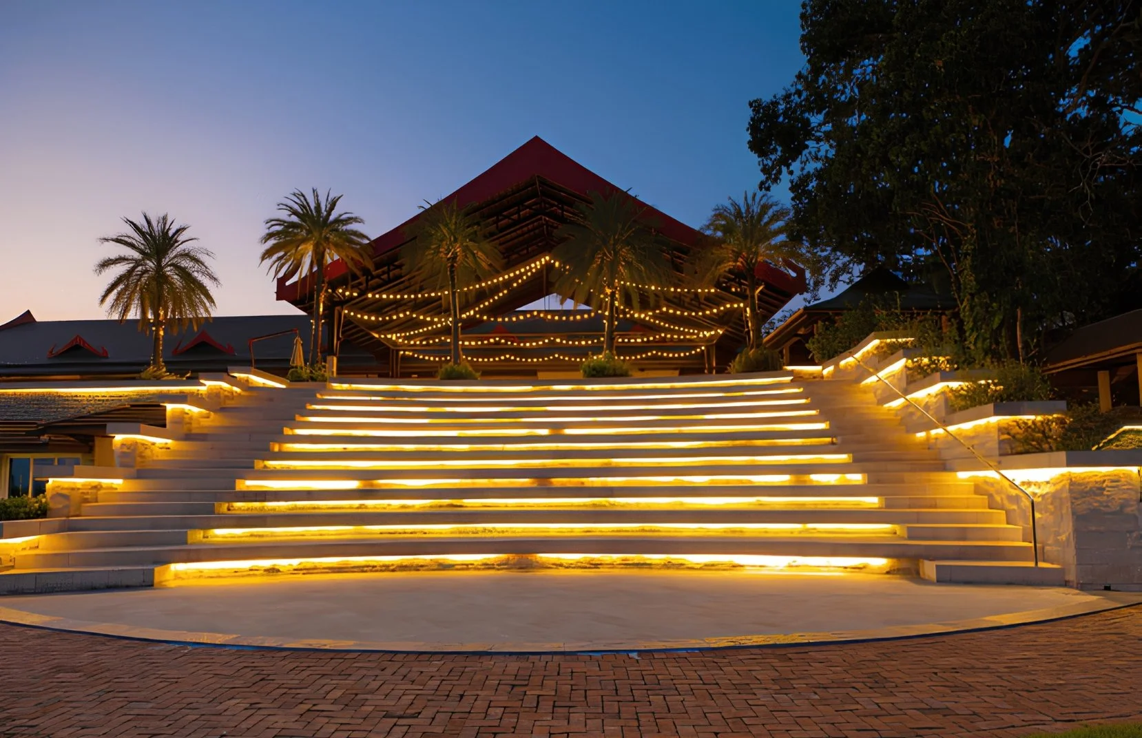 Illuminated staircase leading to an open-air pavilion with string lights, surrounded by palm trees at dusk.
