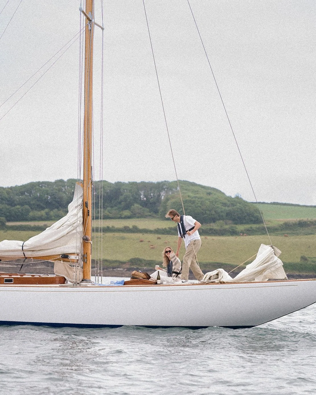 A man and woman on a white sailboat with a wooden mast on a body of water, with green hills and trees in the background. The woman is sitting, wearing sunglasses, and the man is standing, reaching towards her.