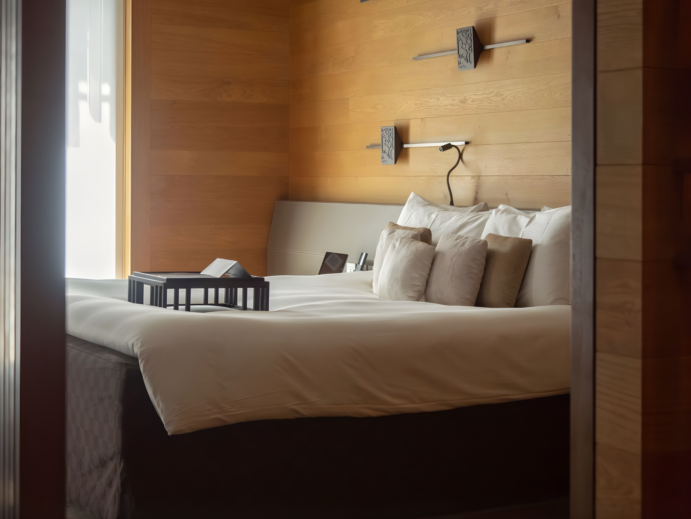View of a hotel room with a bed, pillows, nightstand, lamp, and wooden-paneled wall, taken from outside the room door.