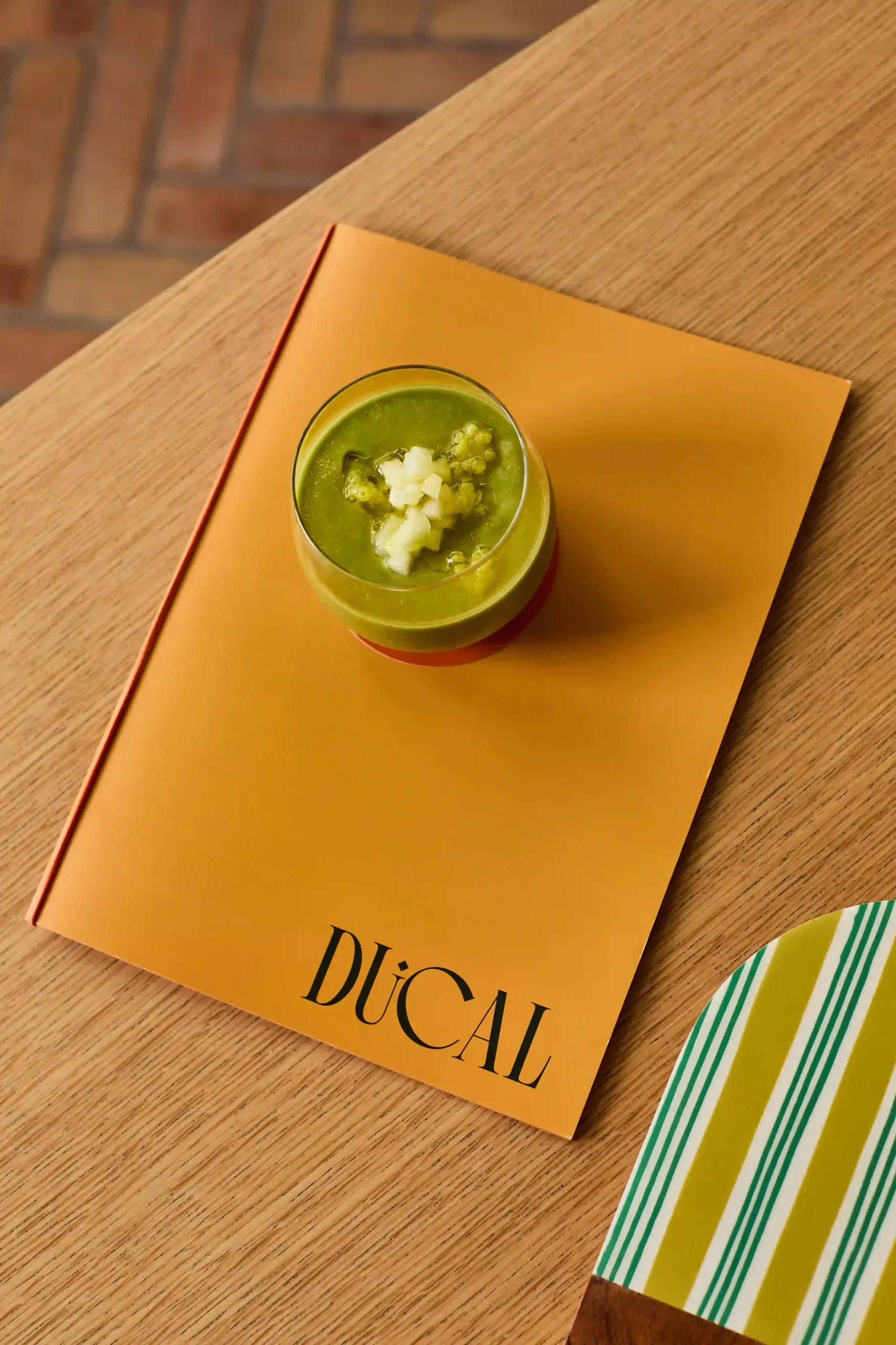 A glass of green liquid with white toppings on a yellow Duocal book on a wooden table.