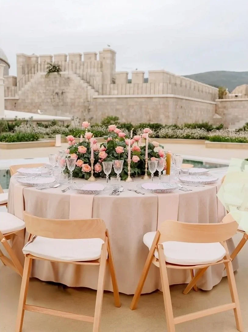 Elegant outdoor dining table with pink flowers and candles, set for a formal event against a historic stone fortress backdrop.