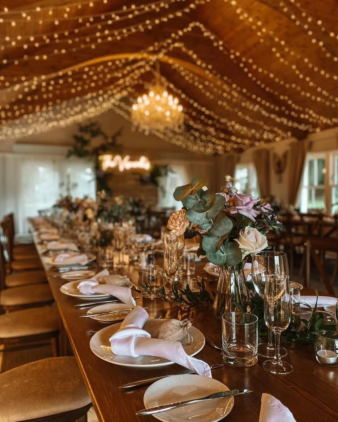 A long wooden dining table set for a celebration with white plates, silverware, napkins, glassware, and floral centerpieces. The ceiling is decorated with string lights and a chandelier, creating a warm and elegant ambiance in a rustic-style room.