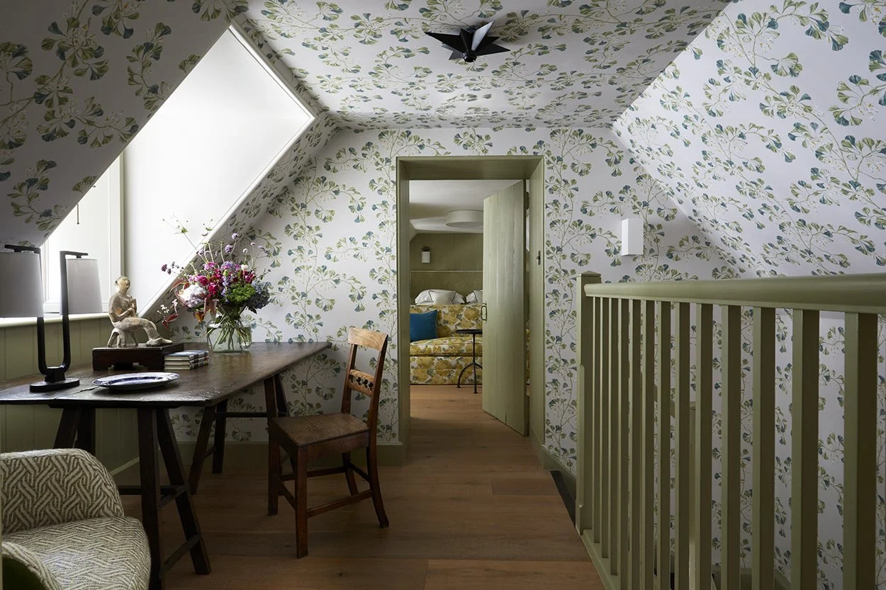 A cozy attic bedroom with floral wallpaper, a wooden desk with a flower arrangement, a zebra figurine, and a chair. A small hallway leads to a second bedroom with a floral-patterned bed and blue cushion.