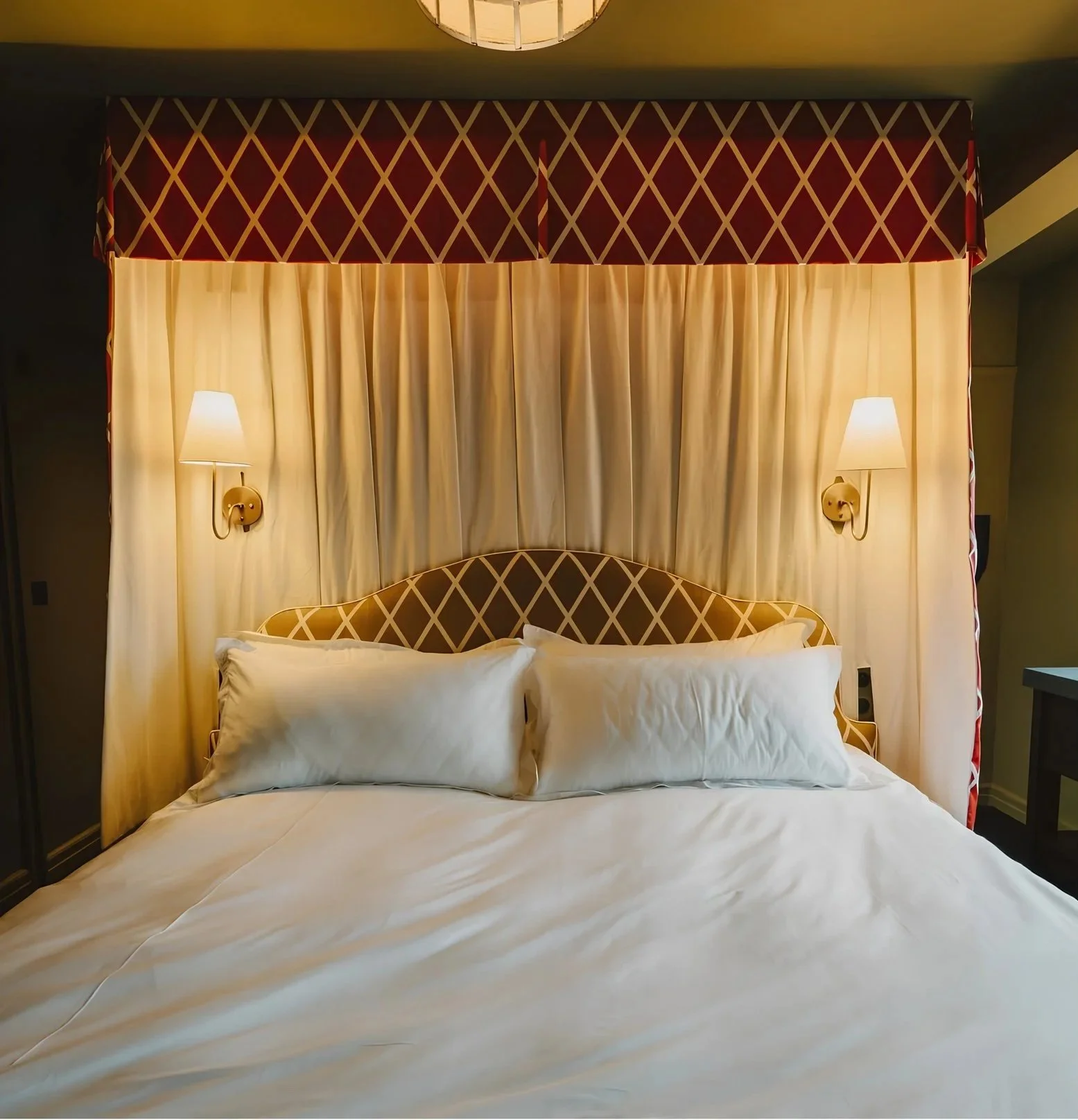 Hotel room bed with a marble headboard, beige curtains behind, two wall-mounted lamps on either side, and a red and beige patterned valance above.