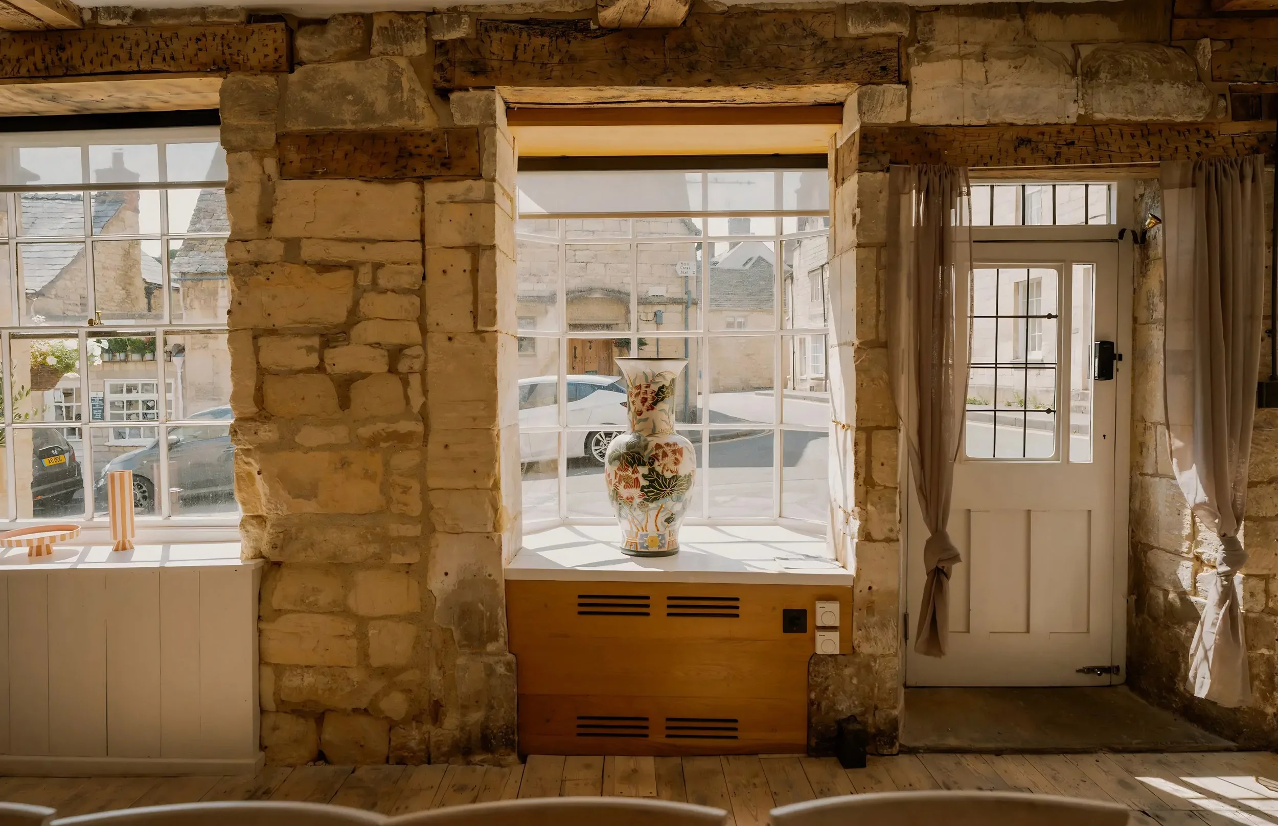 Interior view of a room with large windows, stone walls, a wooden window sill with a decorative vase, and a door with sheer curtains.