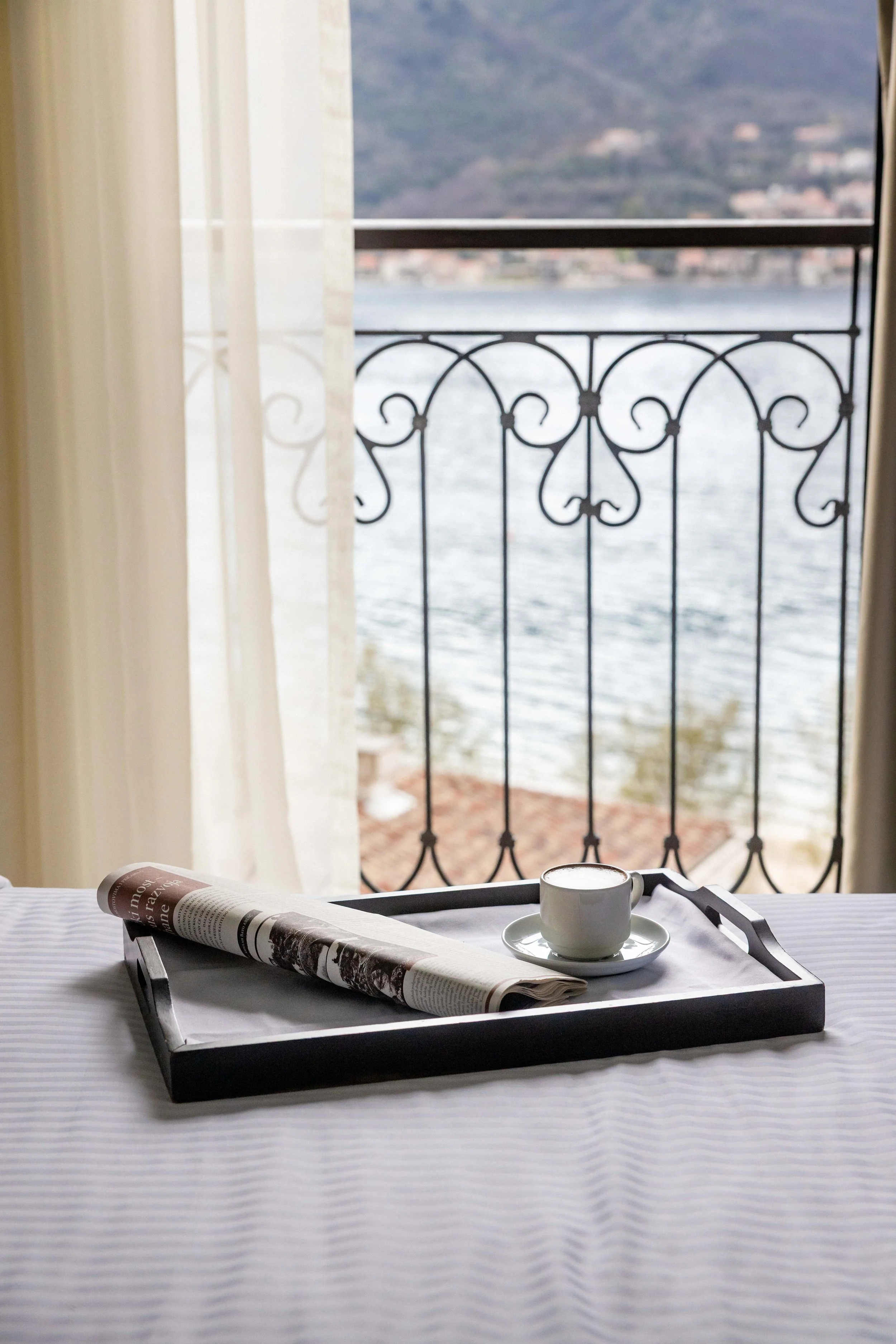 A breakfast tray with a newspaper and a cup of coffee sits on a table covered with a white tablecloth. A view of a lake and distant mountains is seen through an open window with decorative black wrought iron railing. Light curtains frame the scene.