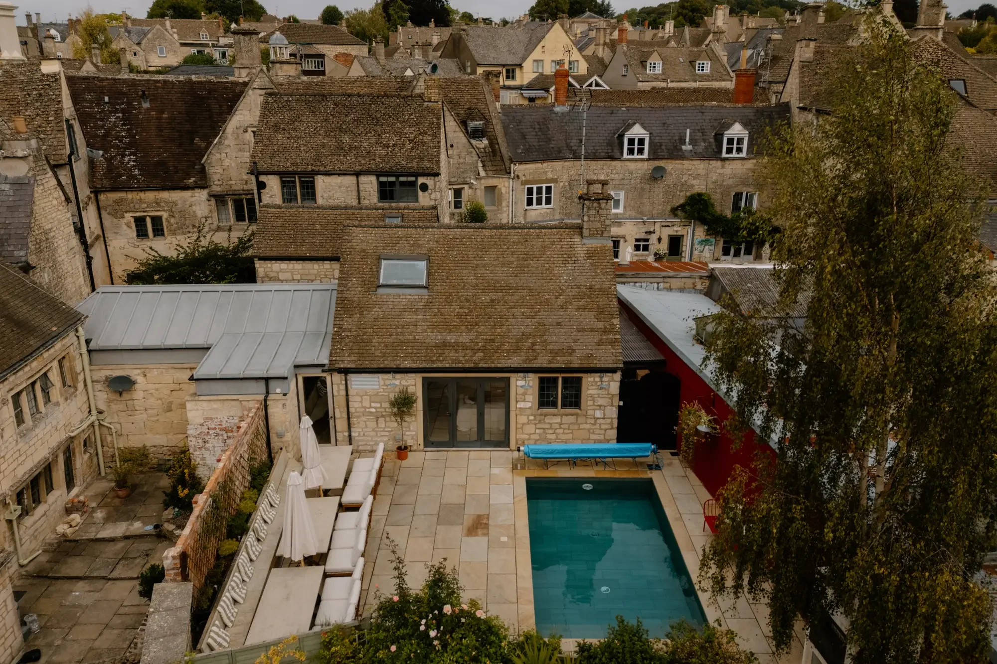 A view of a backyard with a swimming pool, patio furniture, umbrellas, and lounge chairs, surrounded by old stone buildings with tiled roofs.