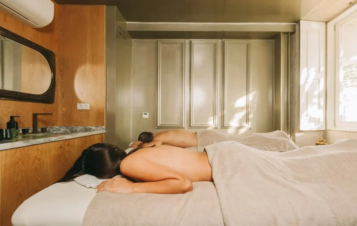 Two people lying face down on massage beds in a spa or massage room, wrapped in towels, with a wooden wall and a window with natural light in the background.