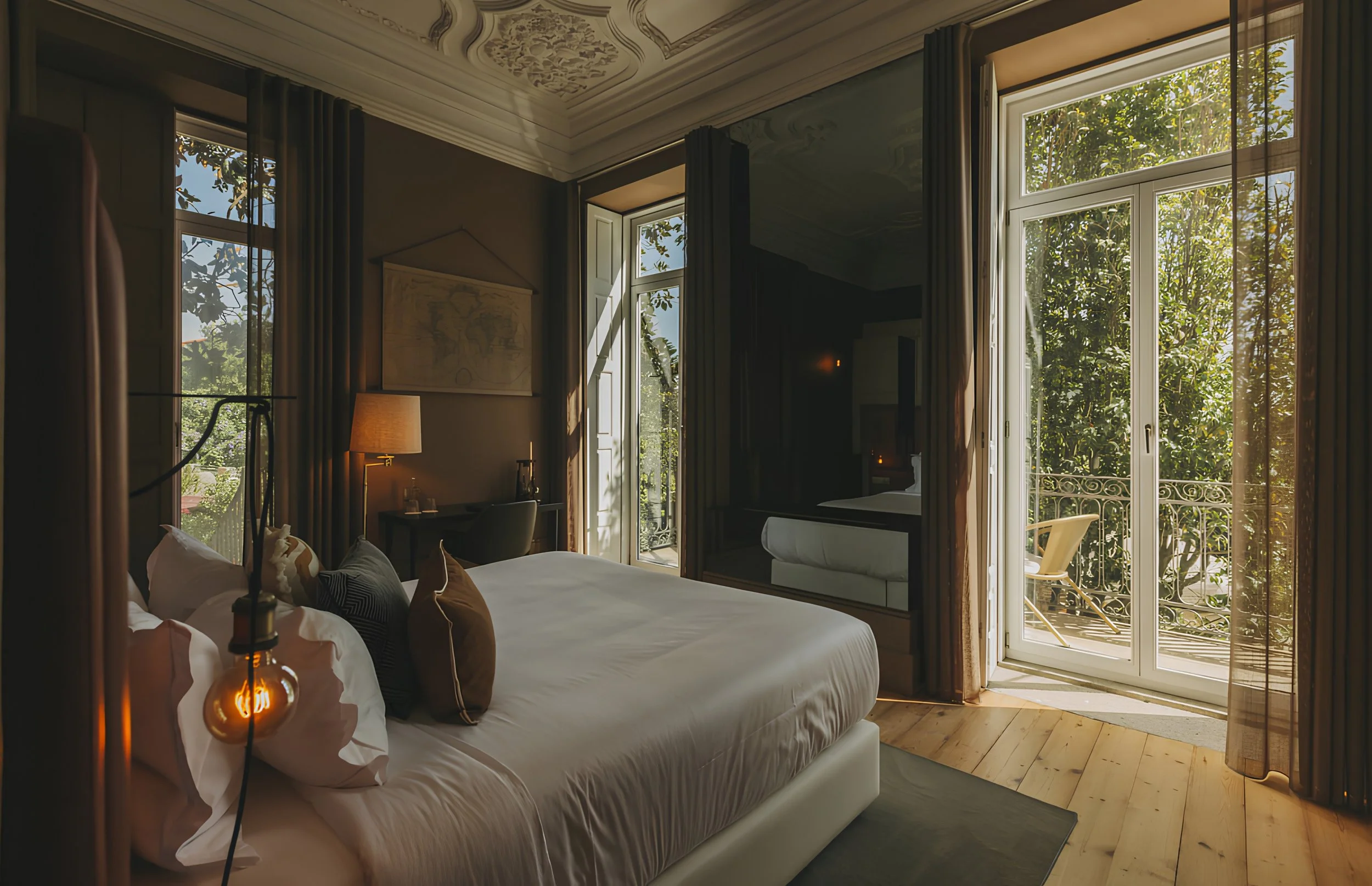 A bedroom with large windows letting in sunlight, with a view of trees outside. The bed has white linens and multiple pillows. There are curtains and some decorative elements like a lamp, a wall map, and elegant ceiling molding.