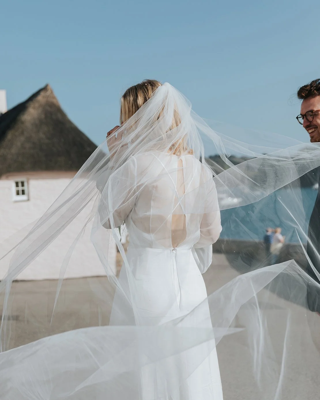 Bride in a white wedding dress and veil on the beach, with a man smiling nearby, under a clear blue sky.