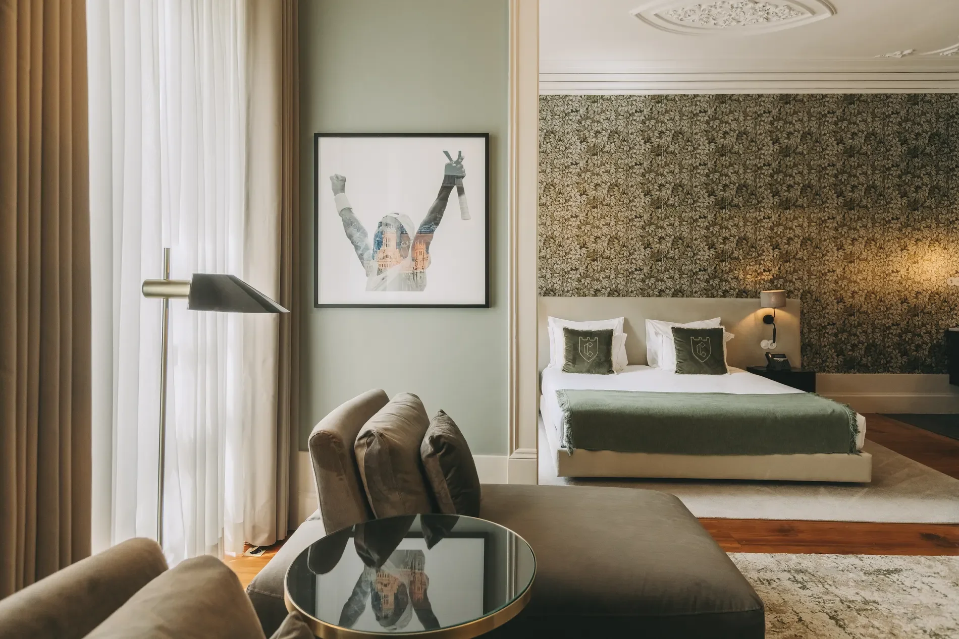 A hotel room with a bed, nightstand, lamp, artwork of a woman with raised arms, and a sitting area with a glass-topped table and cushioned chairs. Light filters through sheer curtains.