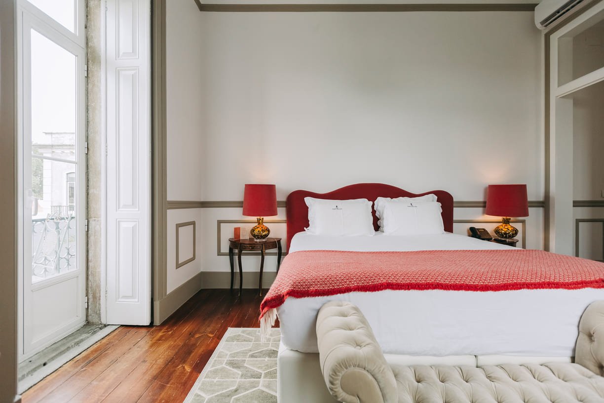 Hotel room with a bed, white sheets, red throw blanket, two white pillows, red bedside lamps, a beige upholstered bench at the foot of the bed, wood flooring, and a balcony door.