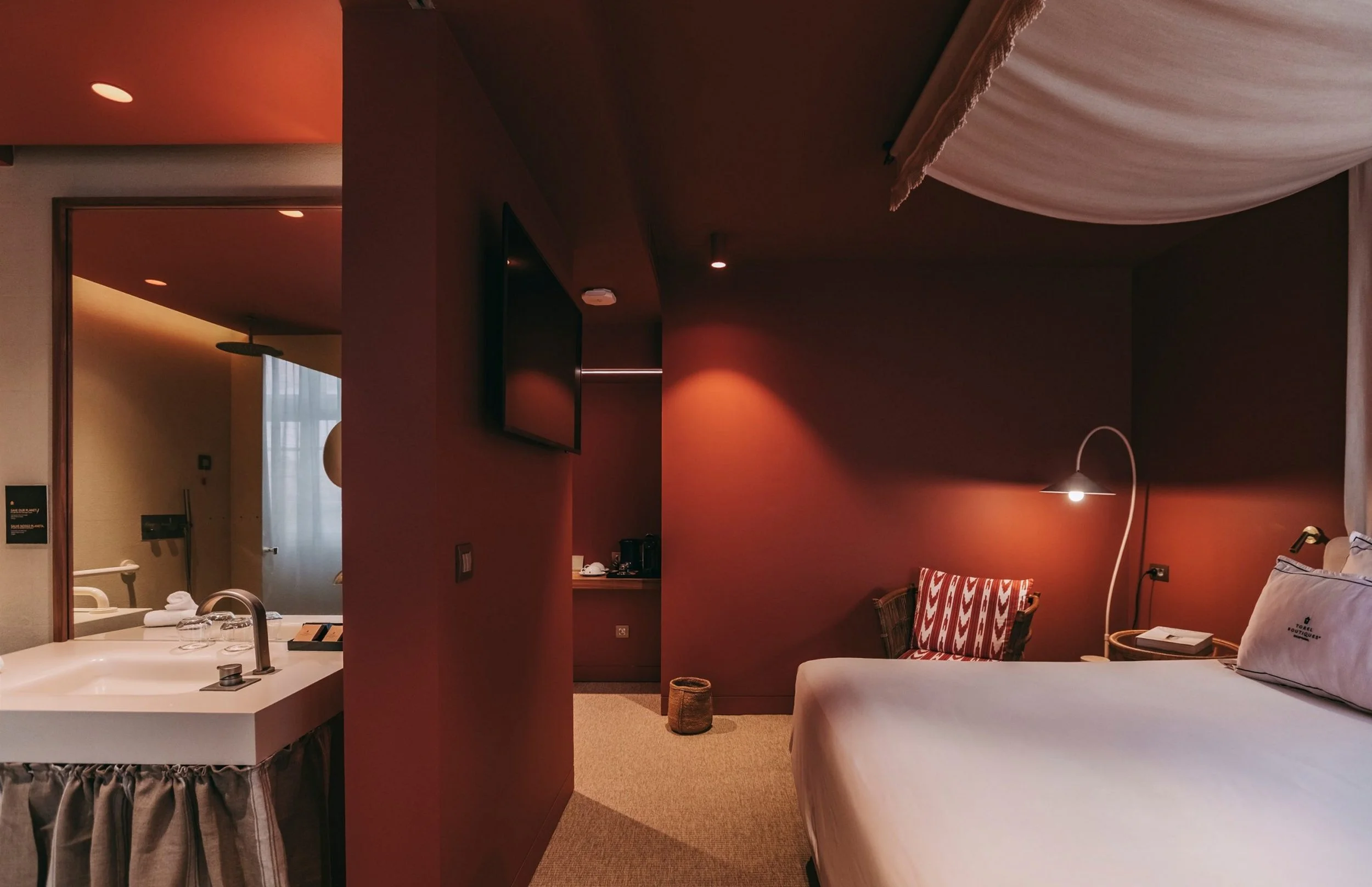 A cozy hotel room with a bed, a ladder-style chair with red and white zigzag pattern upholstery, a wall-mounted flat-screen TV, a white lamp, a small basket, and a draped white canopy on the ceiling. The room has warm orange and soft beige lighting.