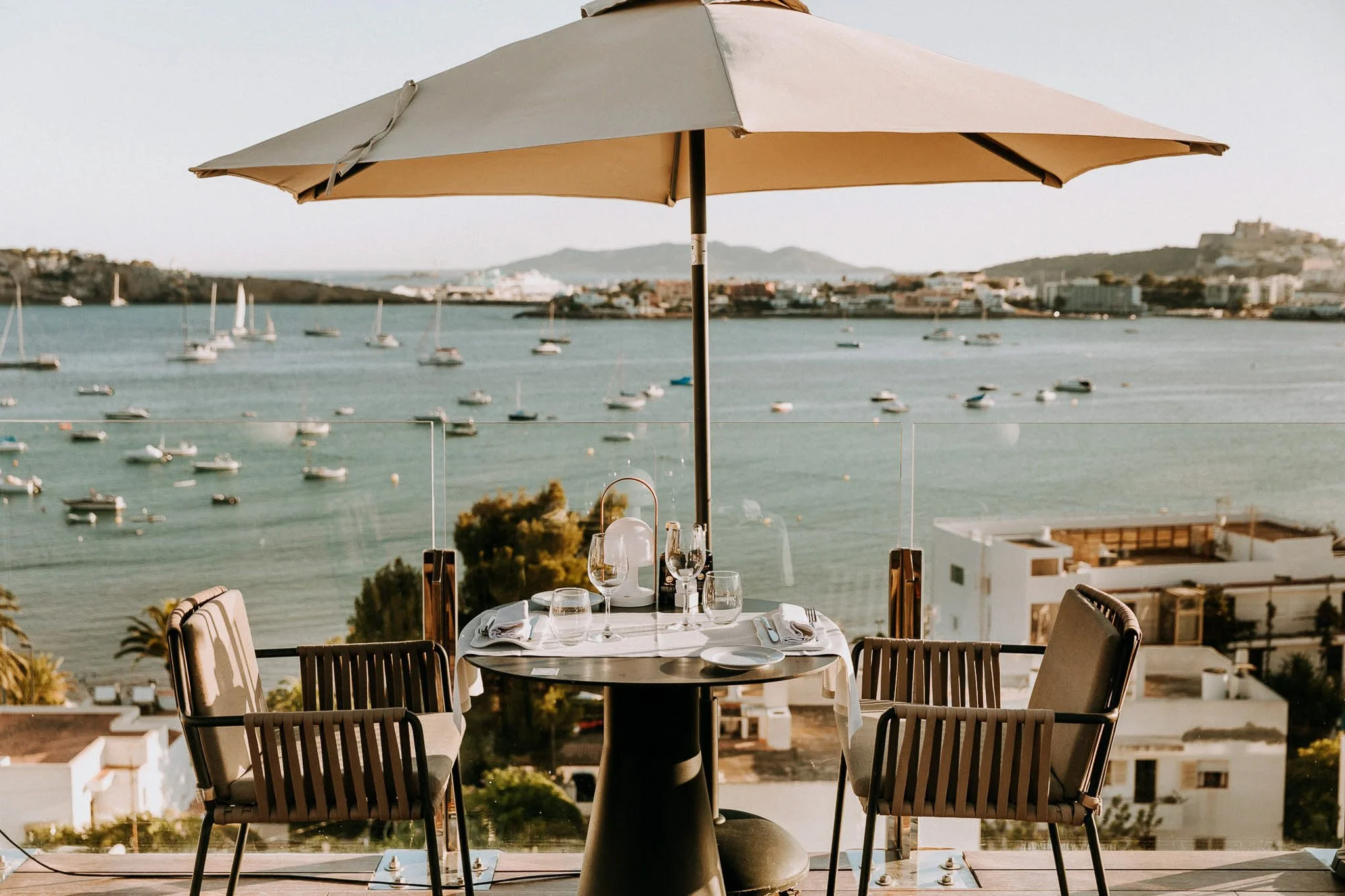 Outdoor dining table set for four with wine glasses, plates, and napkins, under a large beige patio umbrella, overlooking a harbor with numerous sailboats, with buildings and hills in the background.