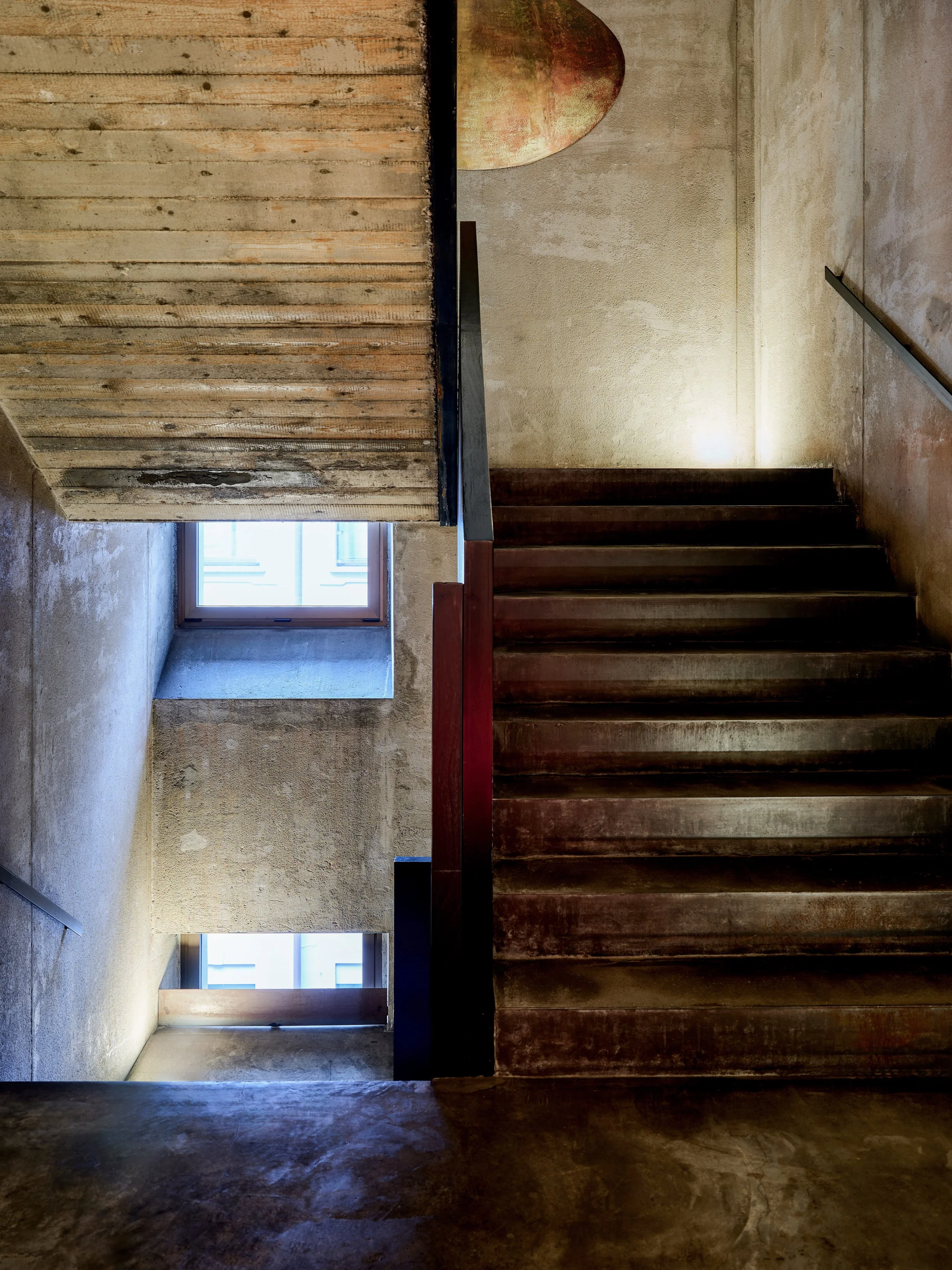 Interior view of a building with a staircase. The left side shows a wall with windows, a countertop and a door, while the right side shows stairs with metal handrails and textured beige walls.