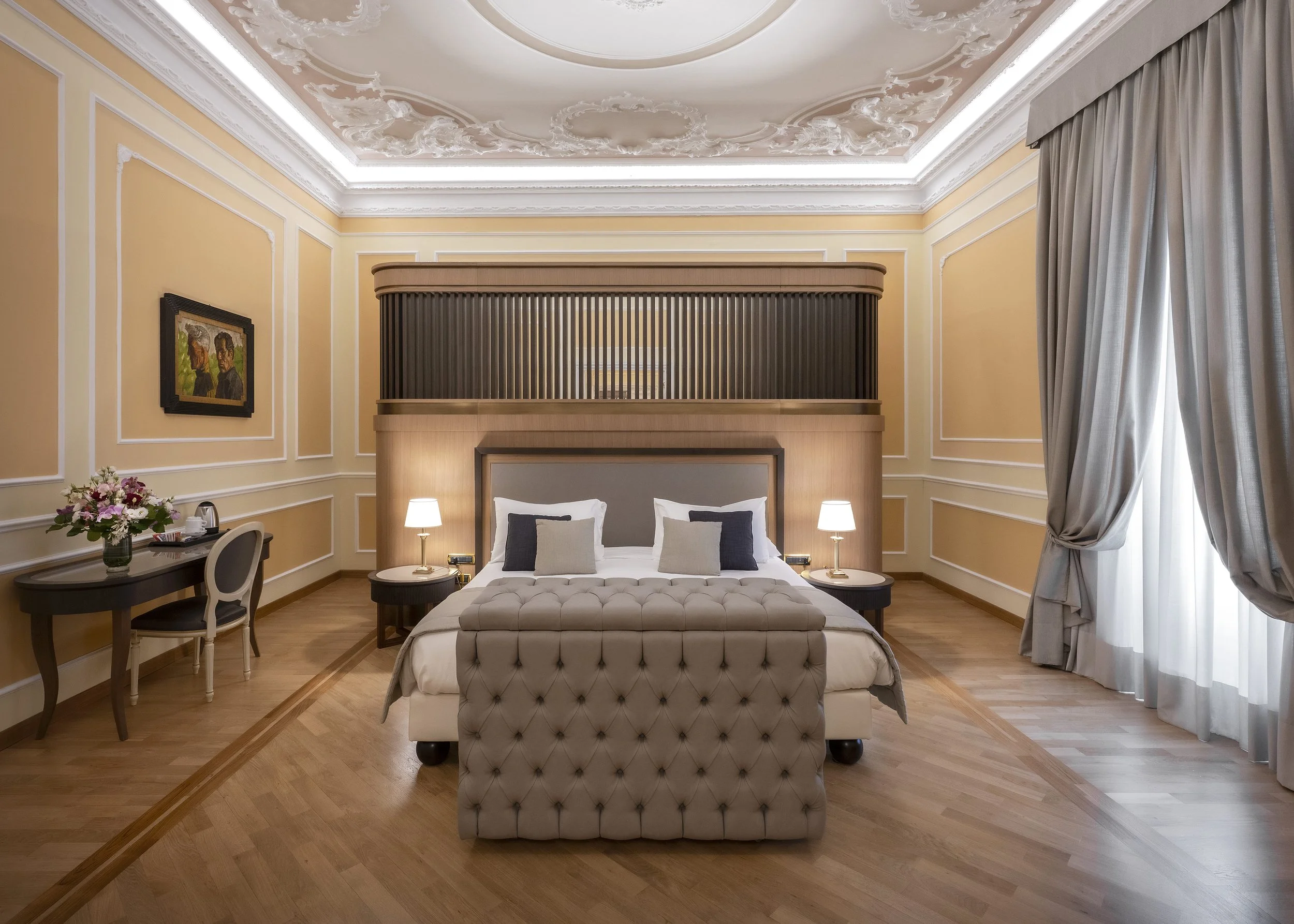 A luxurious hotel room with a large bed, tufted beige bench at the foot, two bedside lamps, a framed painting, a bouquet of flowers, and elegant curtains. The ceiling features ornate white plasterwork and recessed lighting.