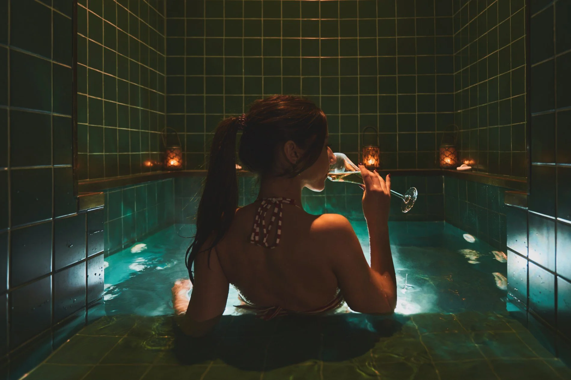 Woman with dark hair in a ponytail, wearing a striped swimsuit, sitting in a tiled hot tub or Jacuzzi, holding a glass of champagne, surrounded by warm candle lighting.