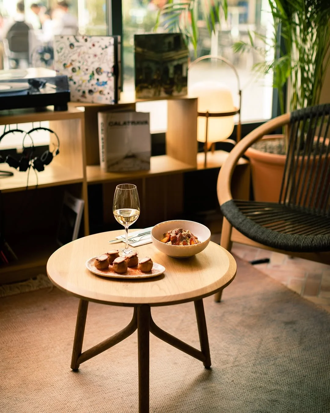 A cozy living room with a small round wooden coffee table holding a glass of white wine, a bowl of food, and a plate of appetizers. In the background, there is a black chair, a shelving unit with LP records and headphones, and a window with green plants.