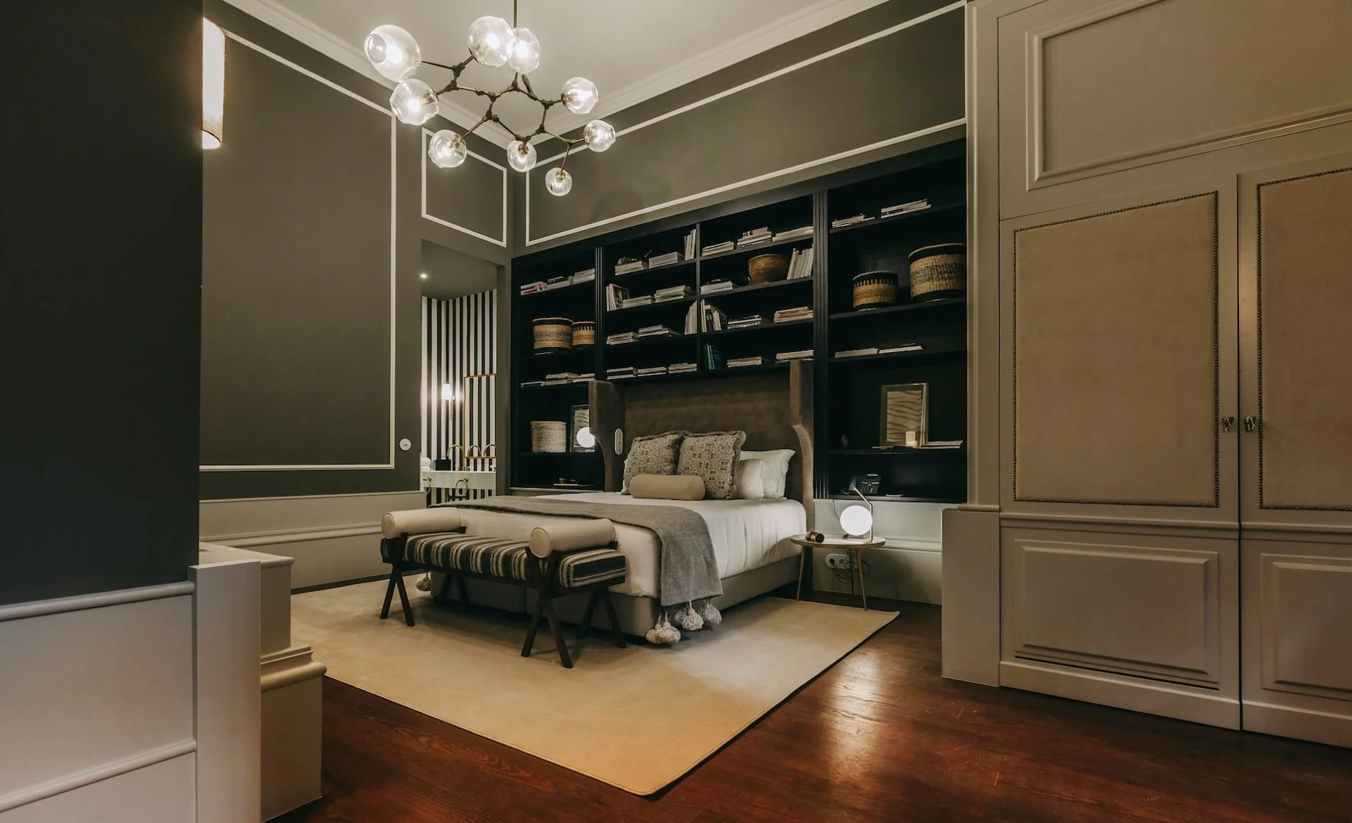 A luxury bedroom with a modern design, featuring a large bed with multiple pillows and a bench at the foot, dark built-in bookshelves behind the bed with decorative baskets and books, a cream-colored area rug, and a stylish chandelier; decor includes