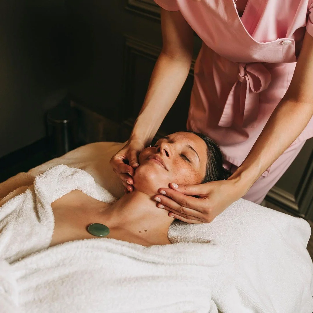 A woman receives a facial massage from a massage therapist in a spa setting, lying on a treatment bed covered with a towel.
