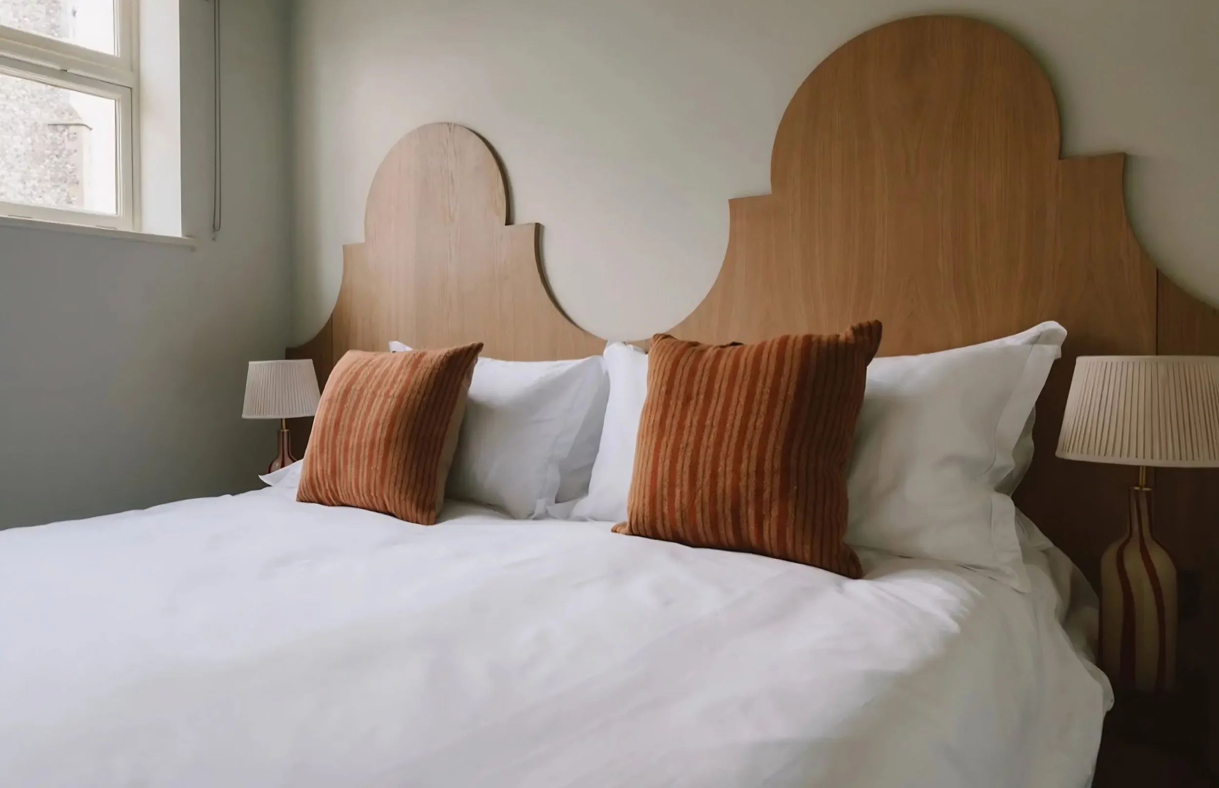 A neatly made bed with white linens and four pillows, two white and two brown striped, in a room with a window and a wooden headboard.