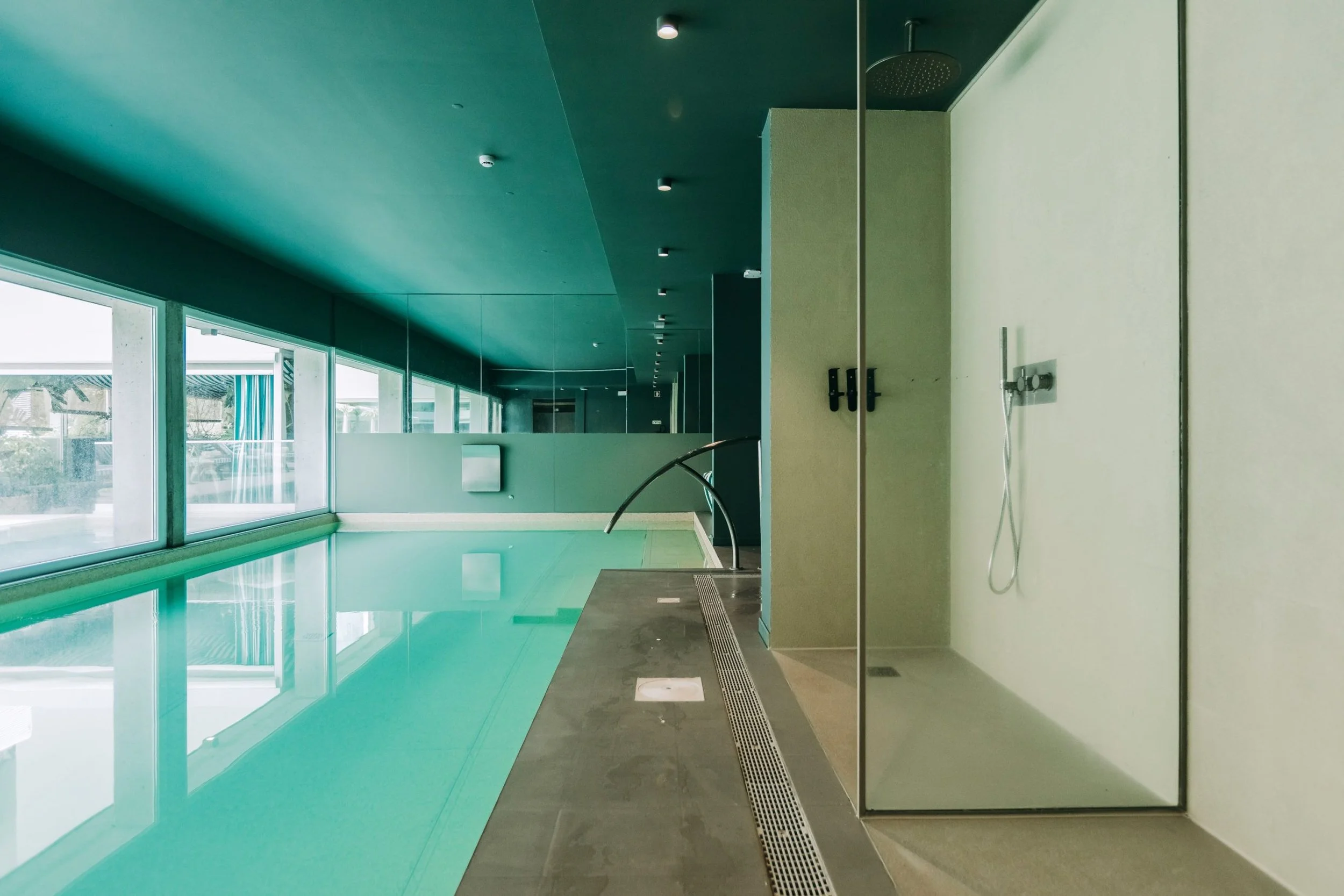 Interior view of an indoor swimming pool area with large windows on one side, green walls, and a modern shower area with a handheld showerhead and drains.