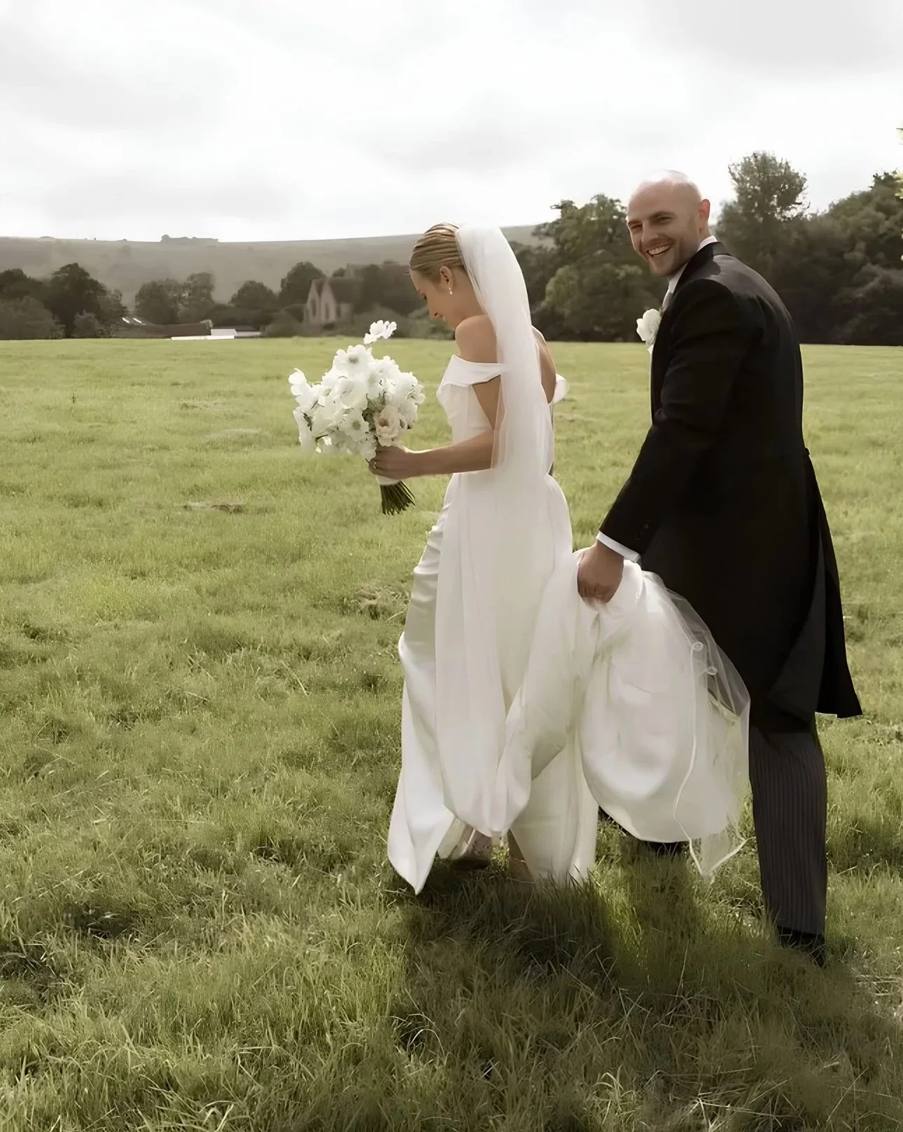 A bride and groom walking in a grassy field on their wedding day, with the groom smiling and the bride holding a bouquet, helps with wedding photography.