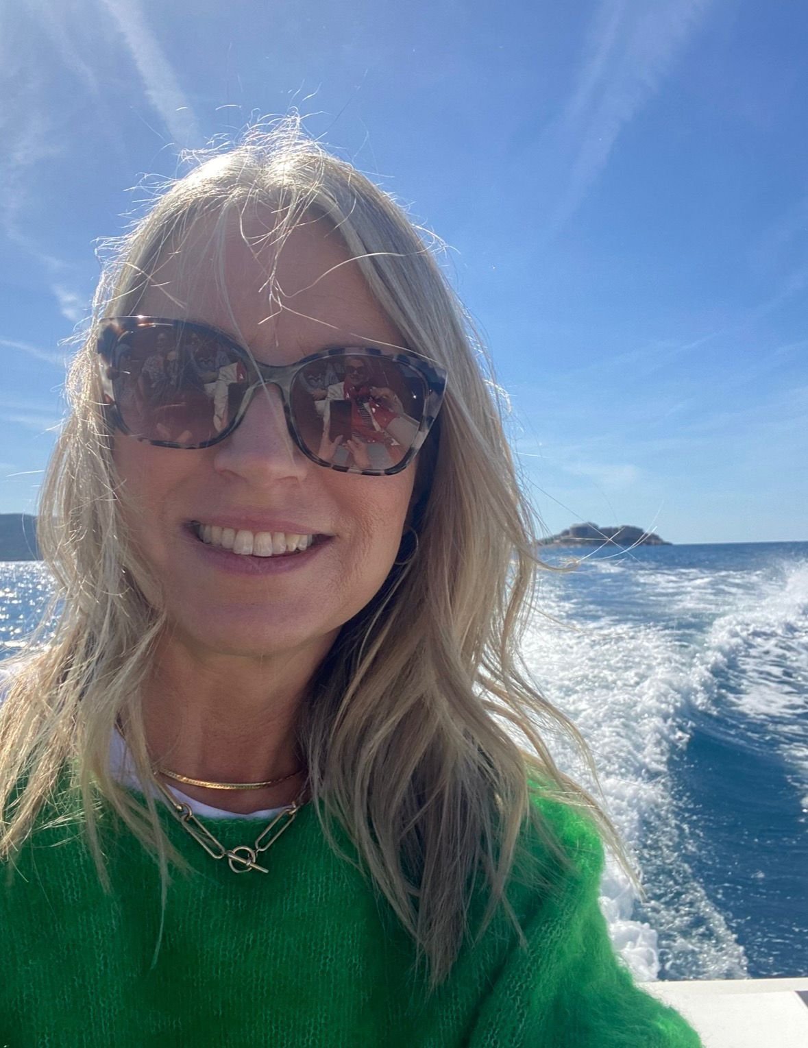 A woman with blonde hair wearing sunglasses and a green sweater is smiling and taking a selfie on a boat with the ocean and a small island visible in the background.