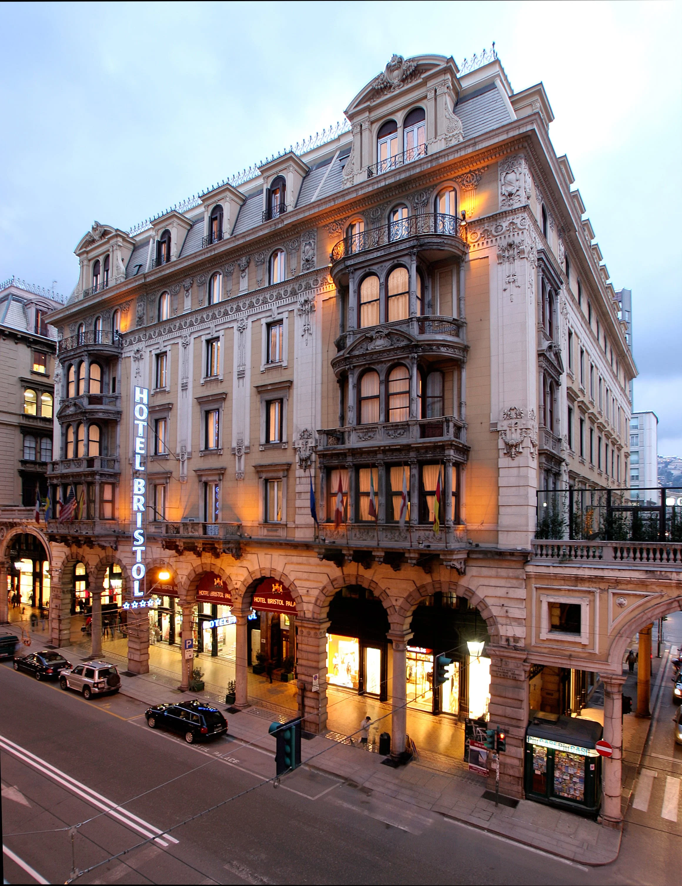 Parisian street scene featuring an elegant historic hotel building with intricate architectural details and balconies, illuminated by warm lights during dusk, with parked cars, traffic lights, and storefronts at street level.