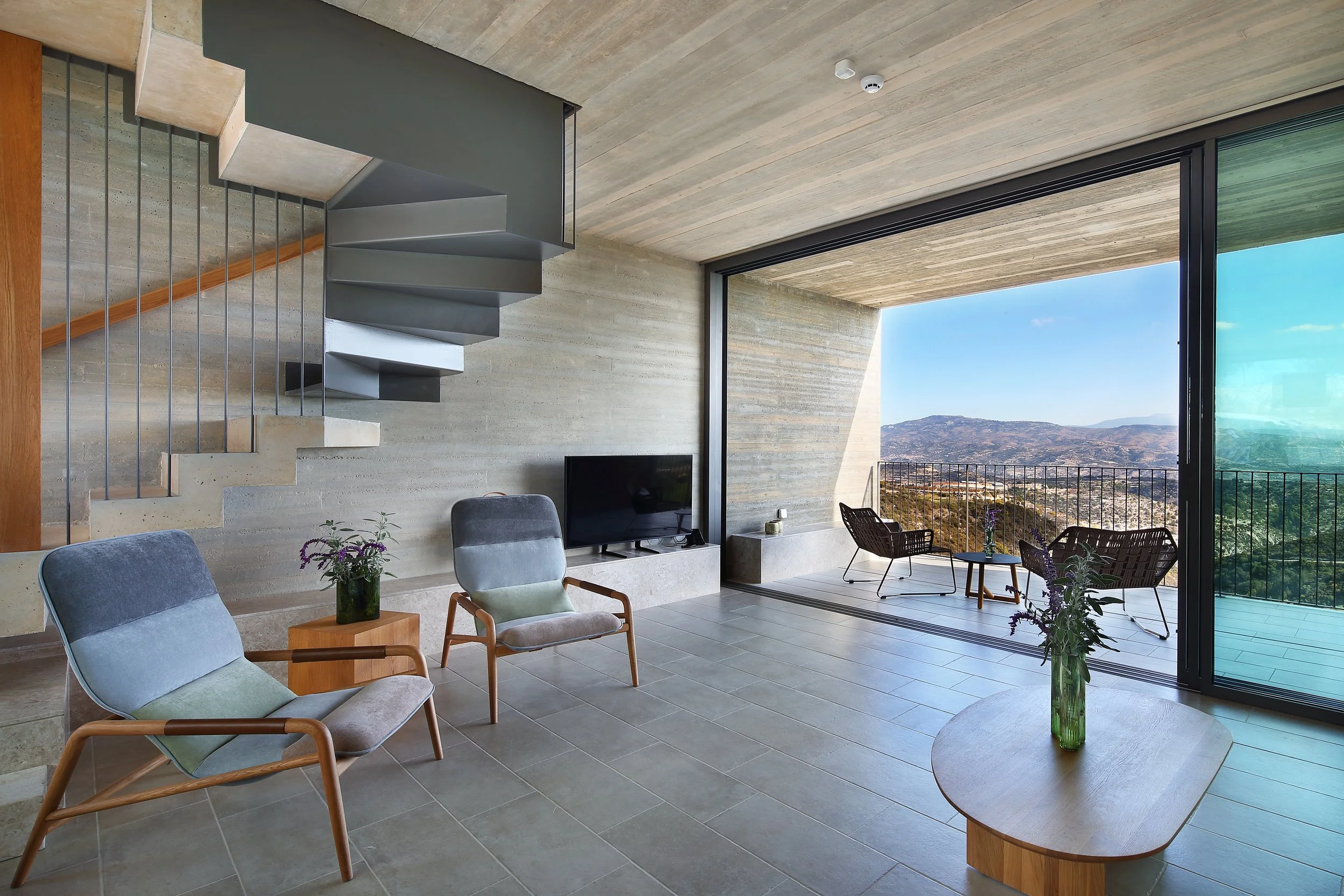 Modern living room with large floor-to-ceiling windows showing a mountain landscape, featuring seating with armchairs, a coffee table with a vase of flowers, a TV, and a staircase with metal railings.