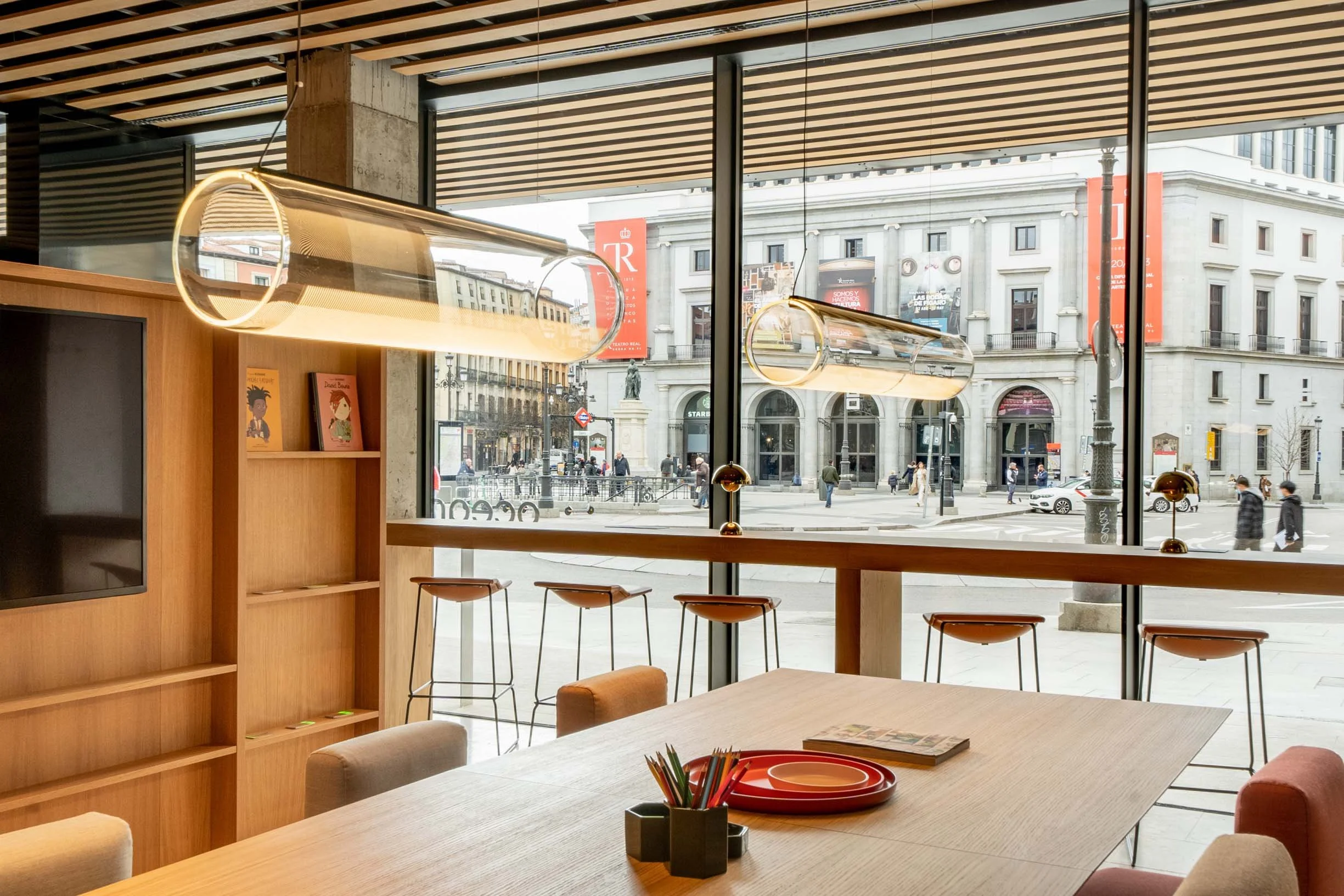 Indoor seating area with a large window showing a city street outside. Inside, there are chairs, a table with colored plates and a container of pencils, and modern hanging light fixtures.