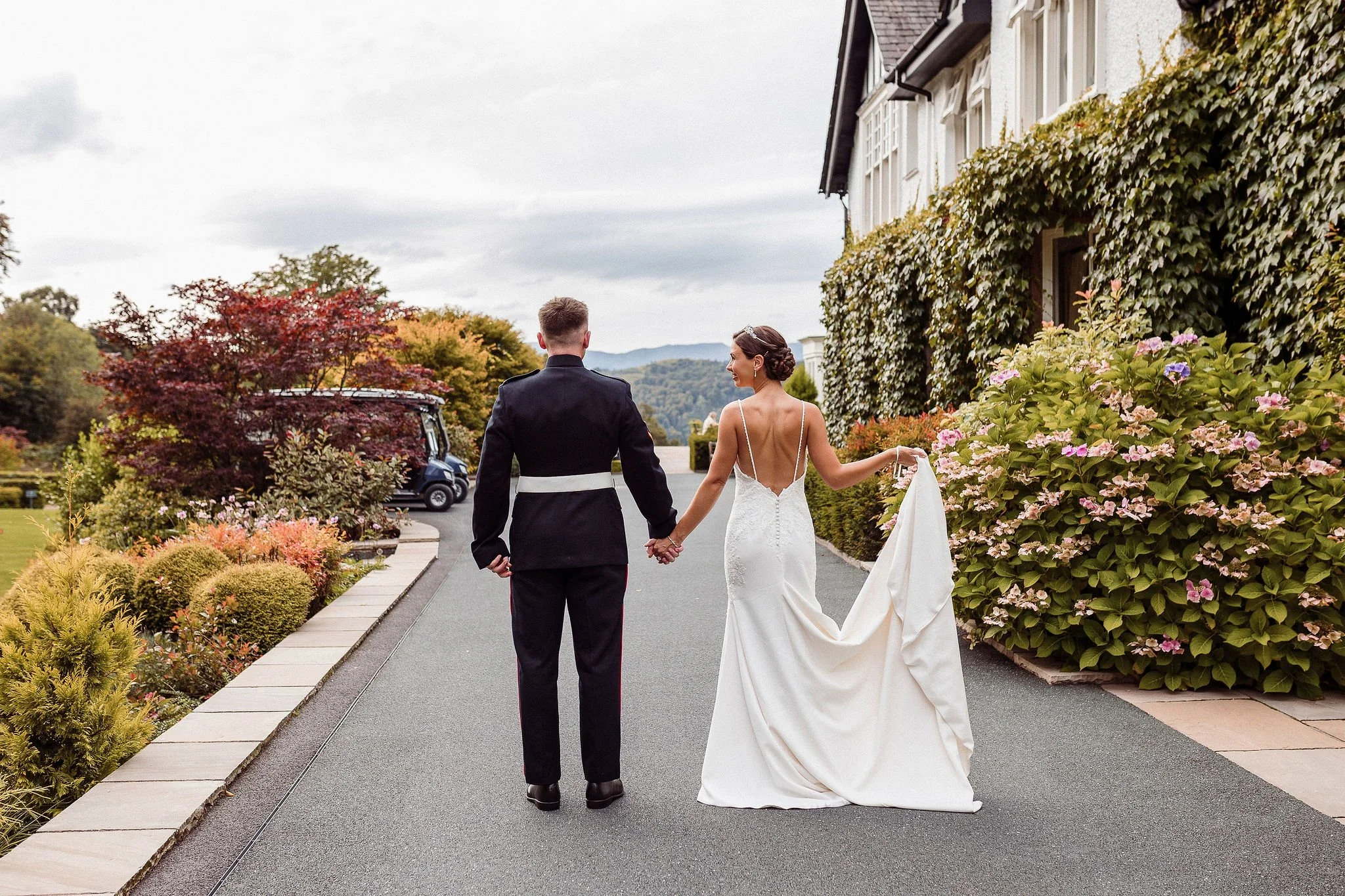 A bride and groom walking hand in hand on a pathway surrounded by colorful autumn foliage, with the bride in a white wedding gown and the groom in a black suit.