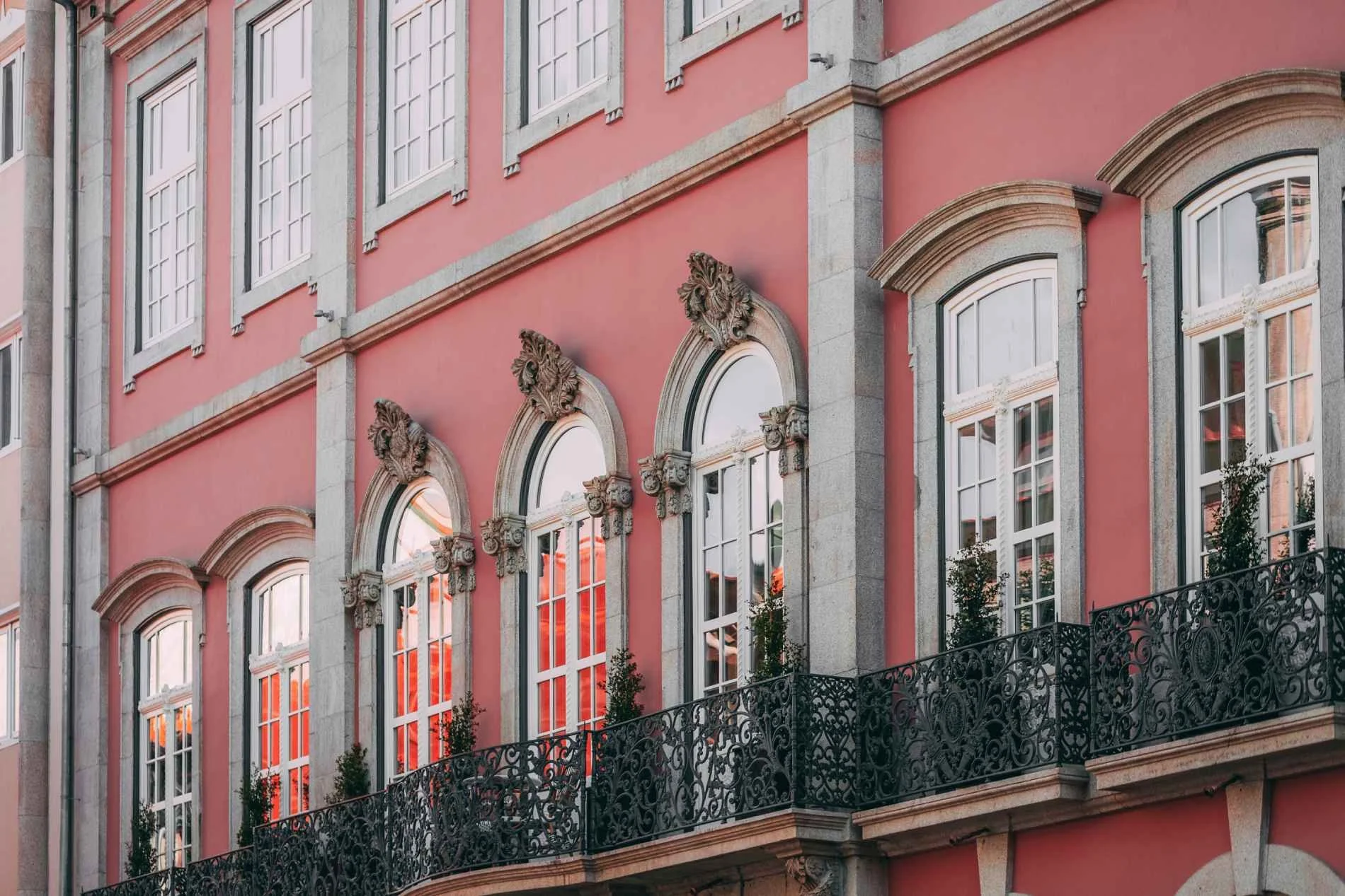 Close-up of a pink historic building with ornate gray stone accents and arched windows, featuring black wrought iron balcony railing and some greenery in front.