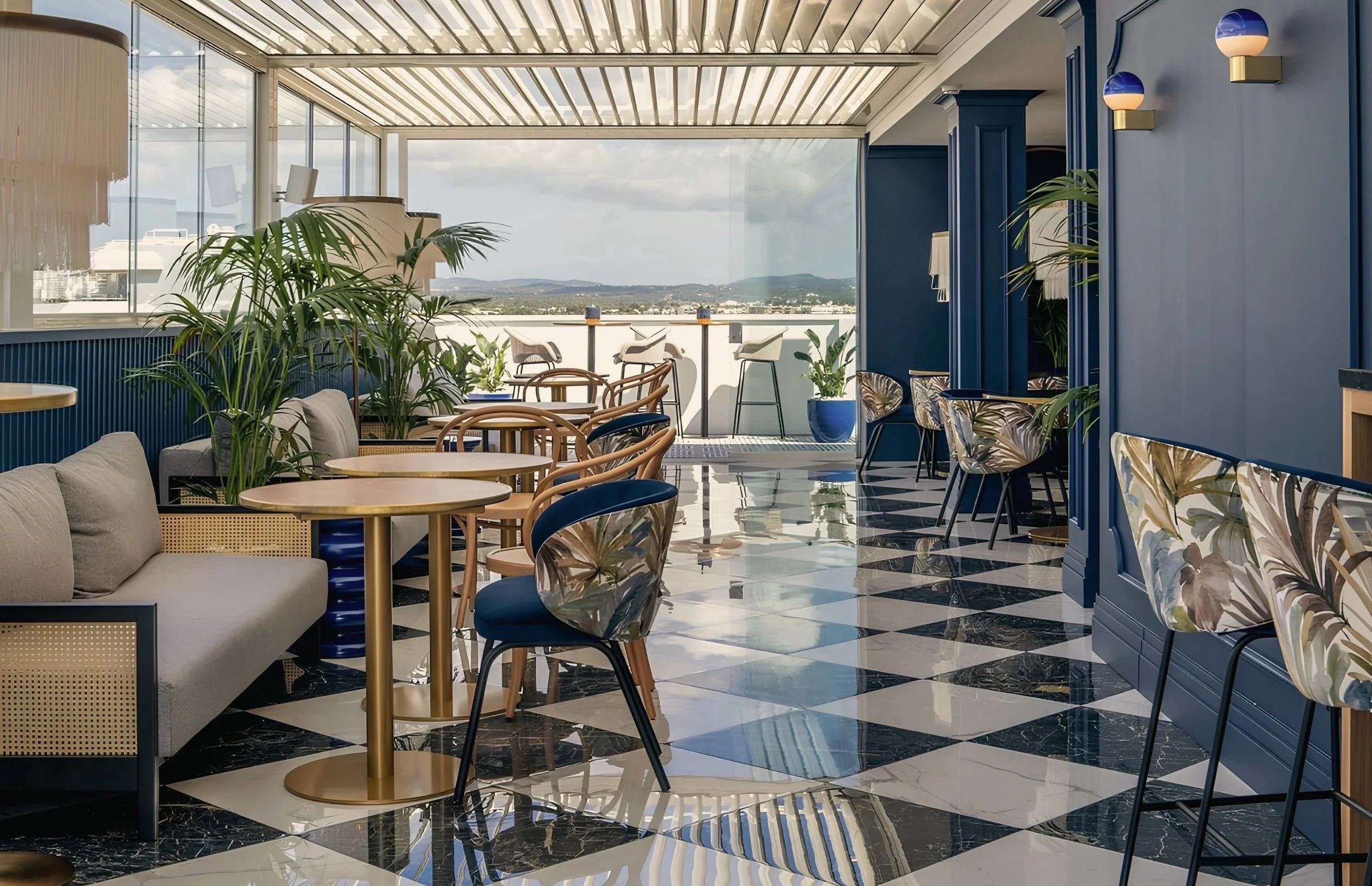 A stylish indoor lounge area with large windows, blue walls, and a view of a cityscape with mountains in the distance. The space features patterned chairs, a sofa, plants, and modern lighting fixtures.