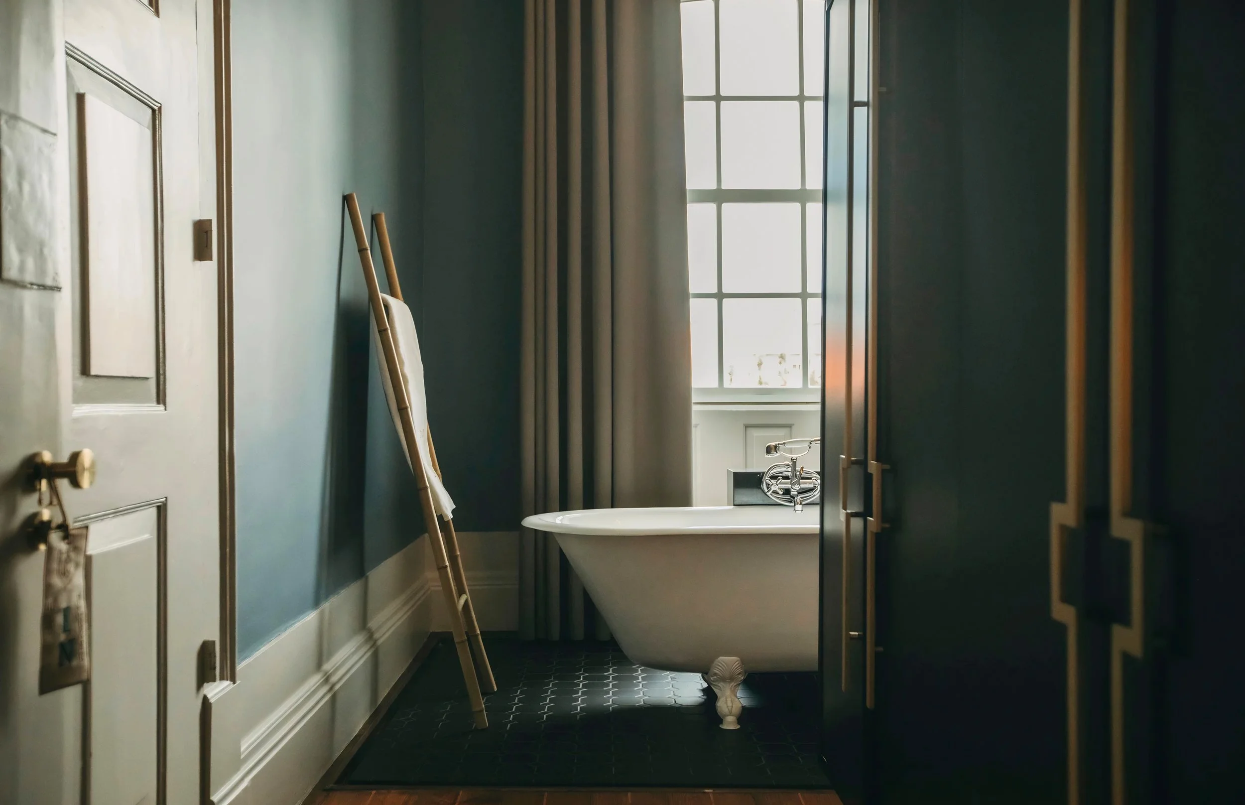 A vintage-style bathroom with a white clawfoot bathtub near a large window with white curtains, a wooden ladder leaning against the wall, and black hexagonal floor tiles.