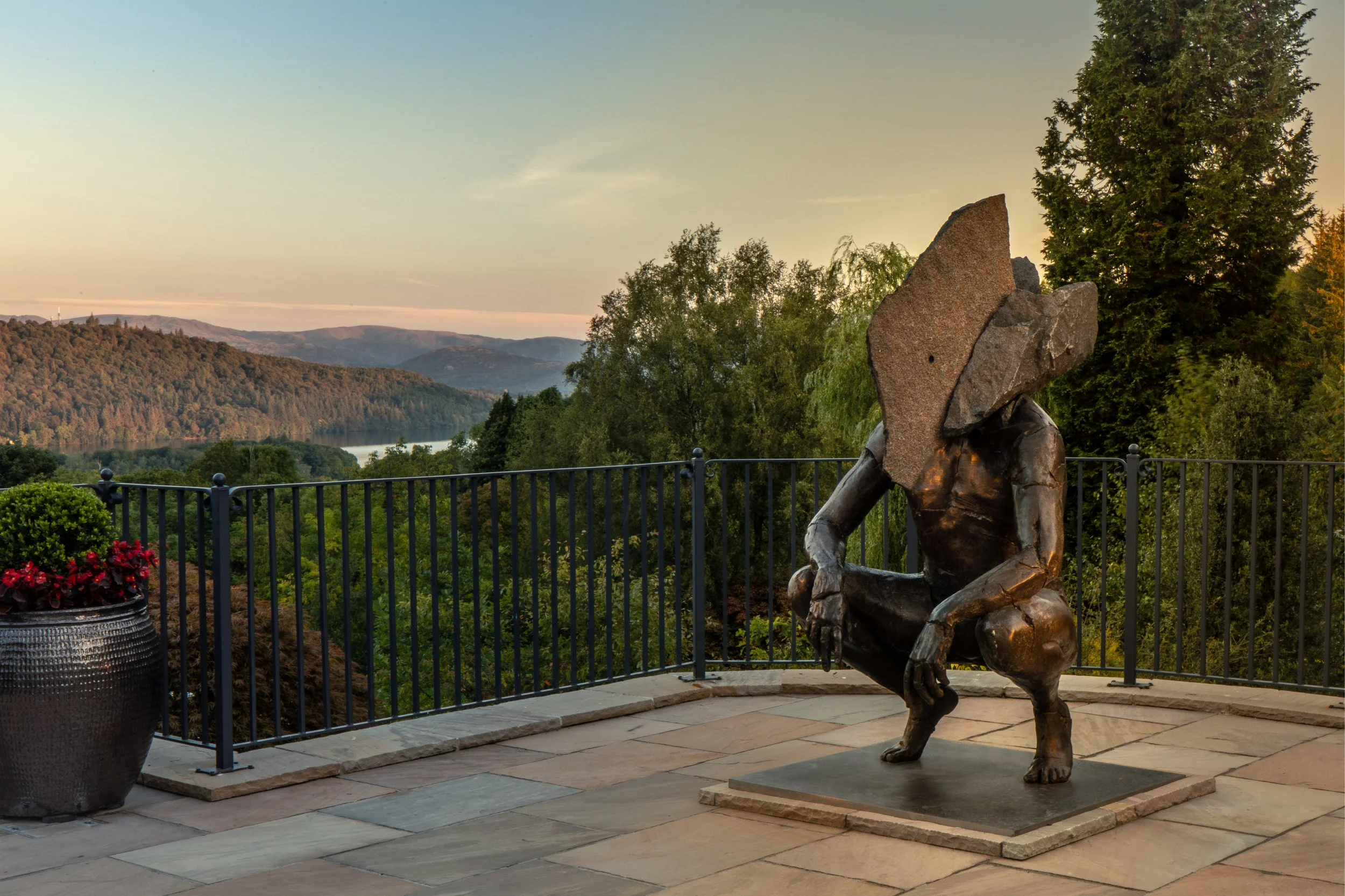 A bronze sculpture of a seated person with their head resting on their knees, located on a patio overlooking a scenic view of hills, trees, and a river at sunset.