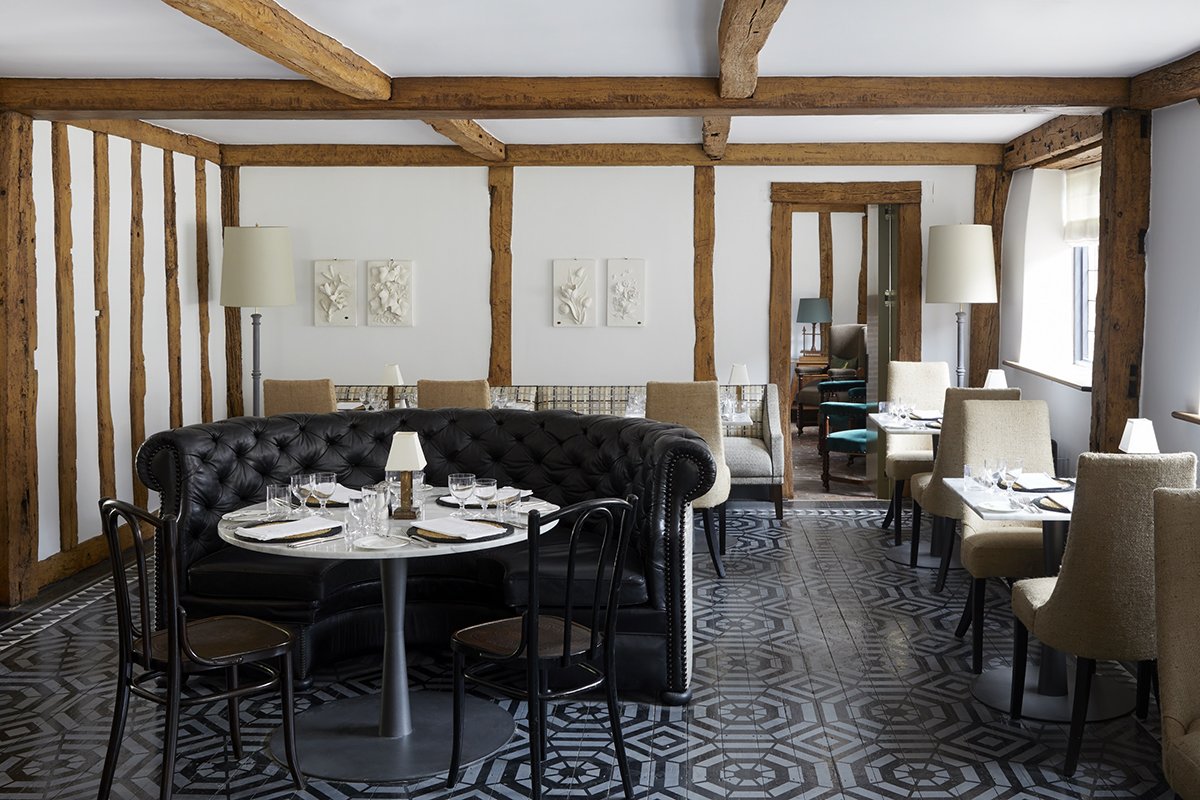 Interior of a restaurant with wooden beams, white walls, and patterned tile flooring. There is a black tufted leather booth around a round table set for dining, with four glasses and plates. Beige and blue upholstered chairs are arranged around other tables, and there are white lamps on the tables and two large floor lamps. Artwork with floral designs is on the walls.