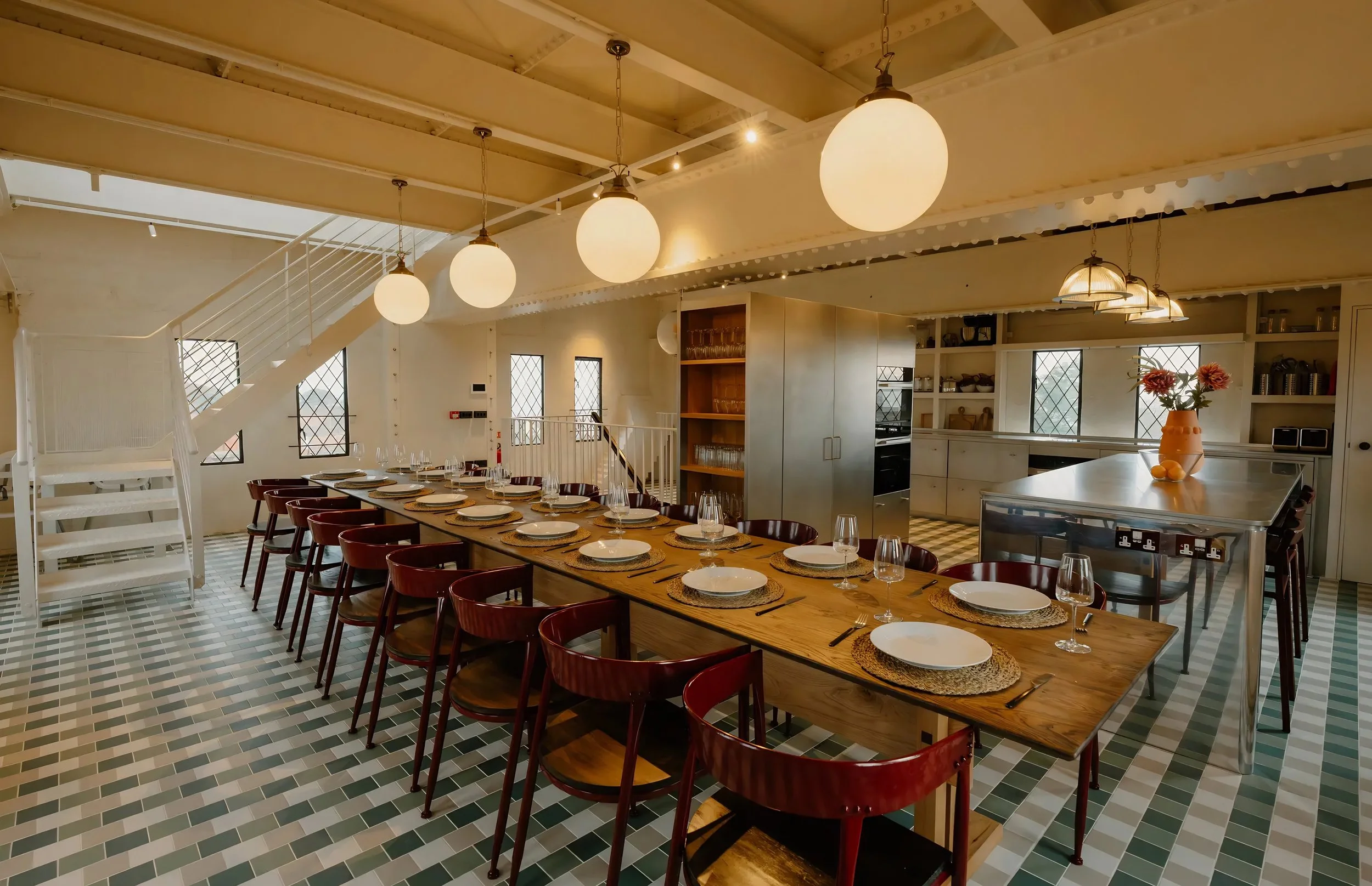Interior of a modern dining room with a long wooden table set with white plates, wine glasses, and placemats. The room has a high wooden ceiling with globe pendant lights hanging above. There are open shelves with glassware and kitchen items, and win