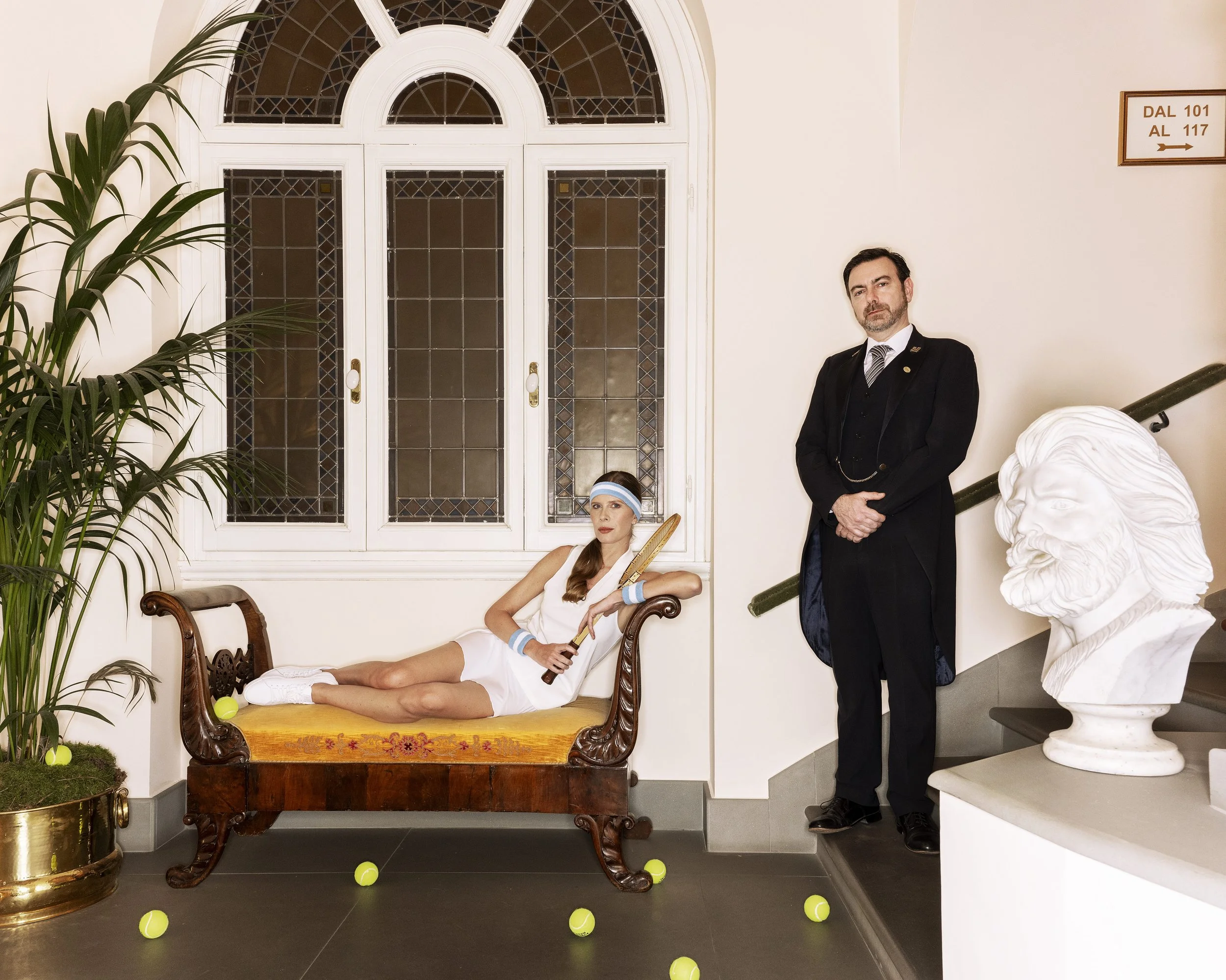 A woman and a man posing indoors near a staircase and a large window with stained glass. The woman is sitting on a vintage wooden bench with tennis balls scattered on the floor around her, holding a tennis racket, wearing a white sports outfit and a 