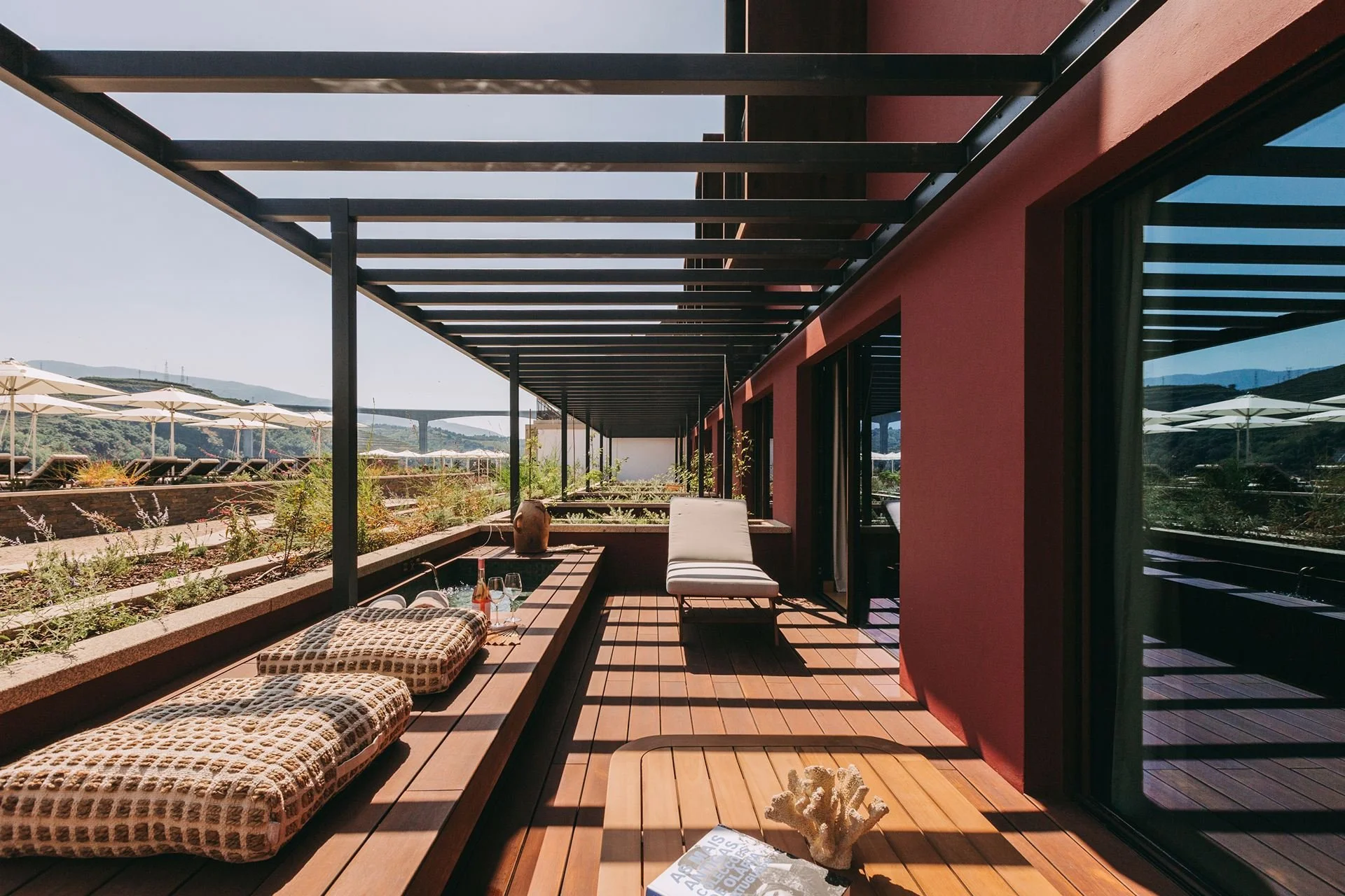 Balcony with wooden flooring, cushioned seating, and potted plants, overlooking a landscape with hills and umbrellas in the distance.