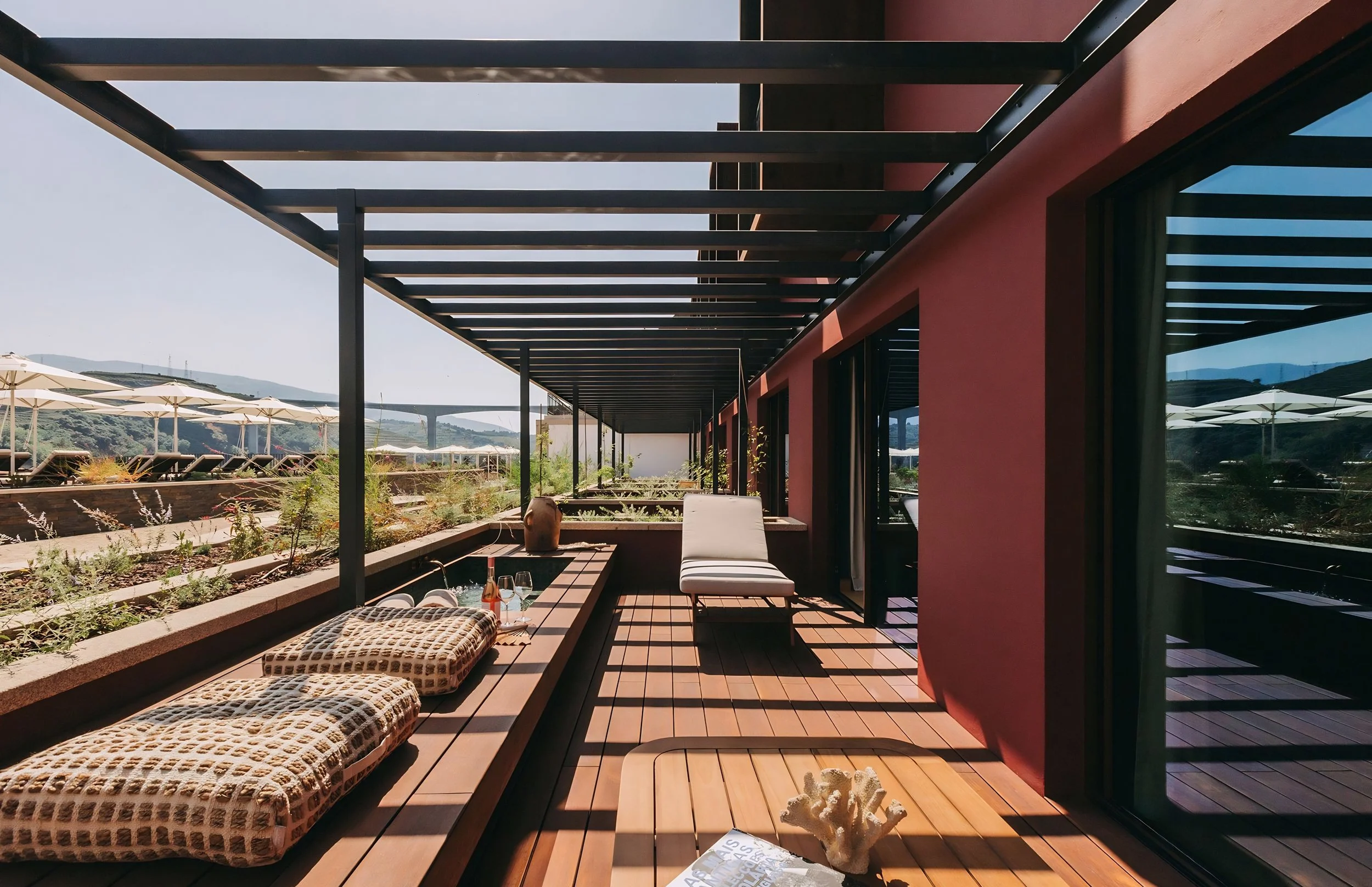A modern outdoor patio with wooden deck, lounge chairs, potted plants, and a red building with large glass windows. There is a black metal pergola overhead and an view of distant hills with umbrellas and lounge chairs in the background.