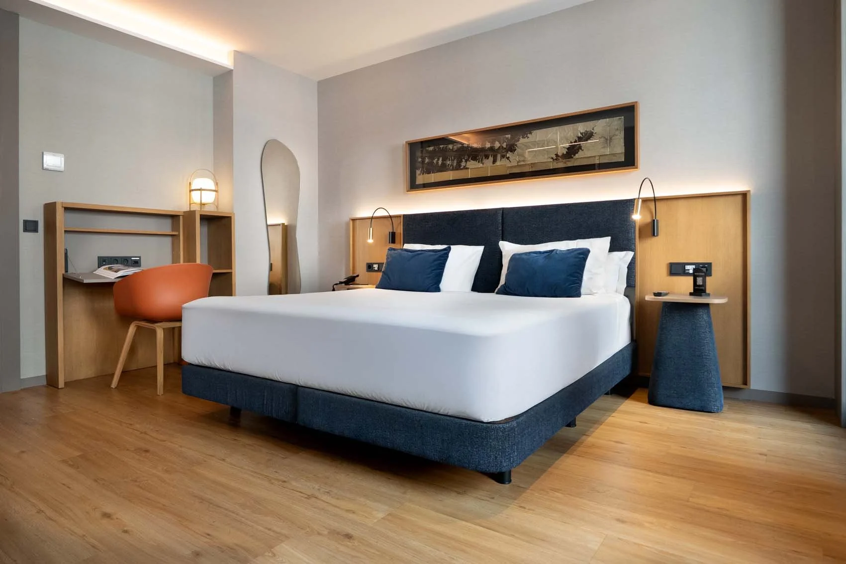 Modern hotel room with a queen-sized bed, navy blue pillows, wooden headboard, wall-mounted reading lights, a small bedside table, a desk with a chair, and minimalist decor.