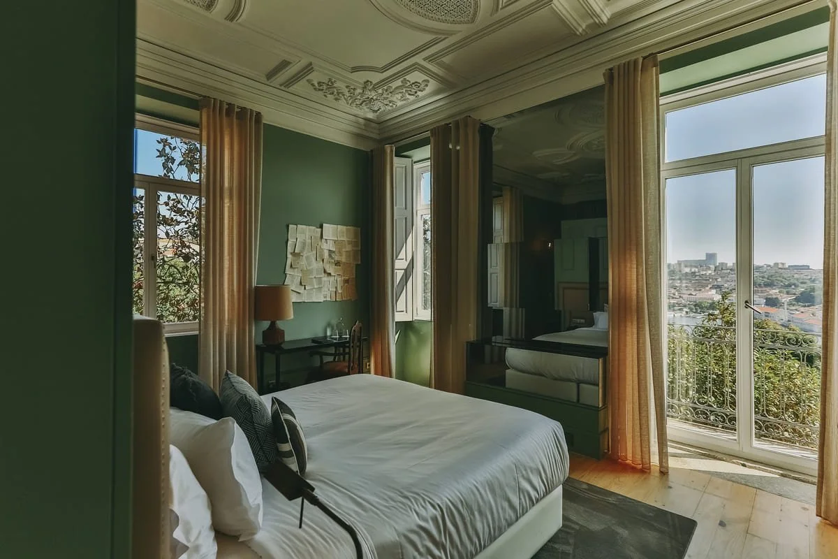 A bedroom with large windows and a balcony overlooking a cityscape, featuring a bed with black and white pillows, a small desk with a lamp, and sheer beige curtains.