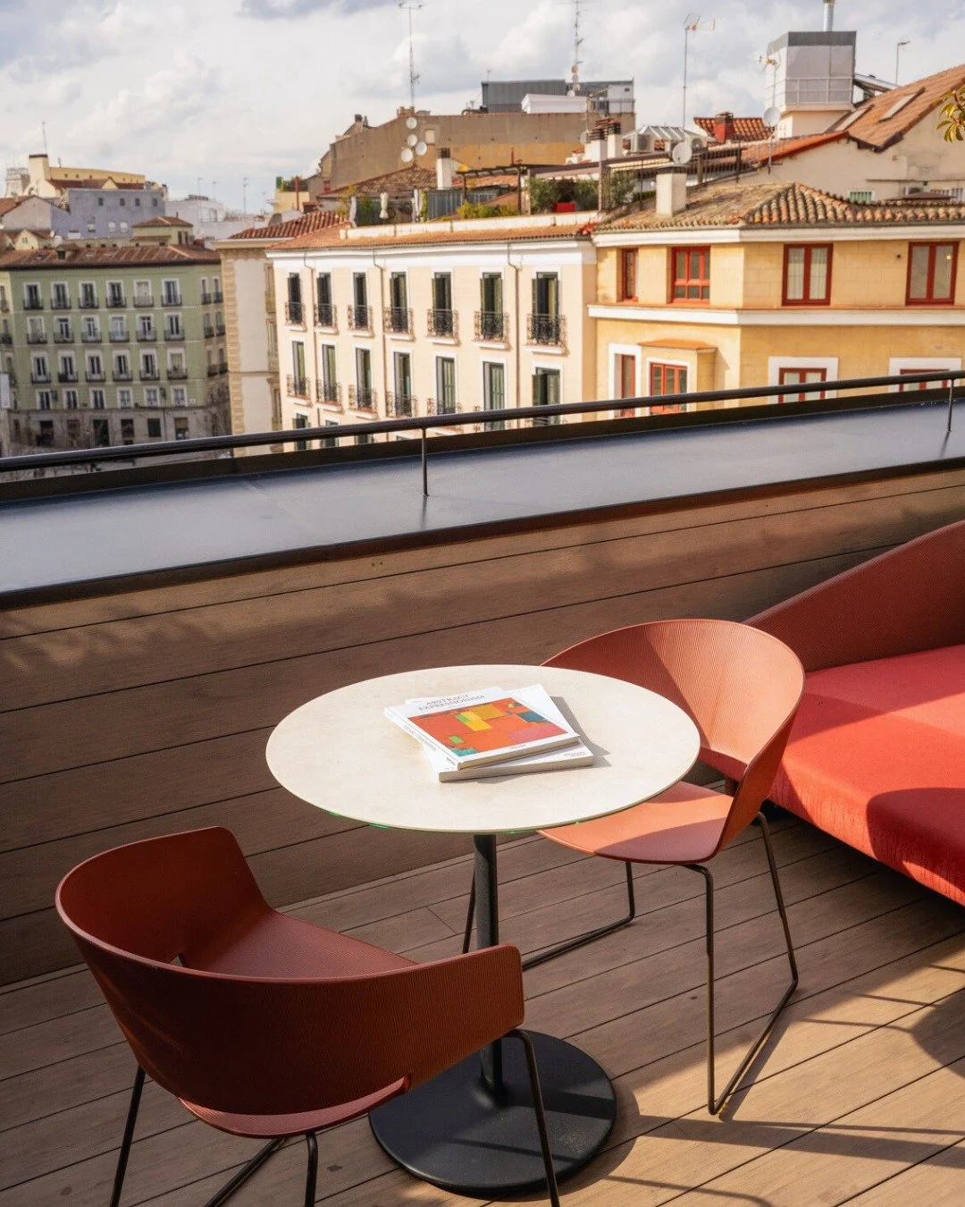 A terrace with a small round table, two pink chairs, and a pink sofa. The table has a magazine on it. In the background, there are colorful buildings with tiled roofs and multiple antennas, under a partly cloudy sky.