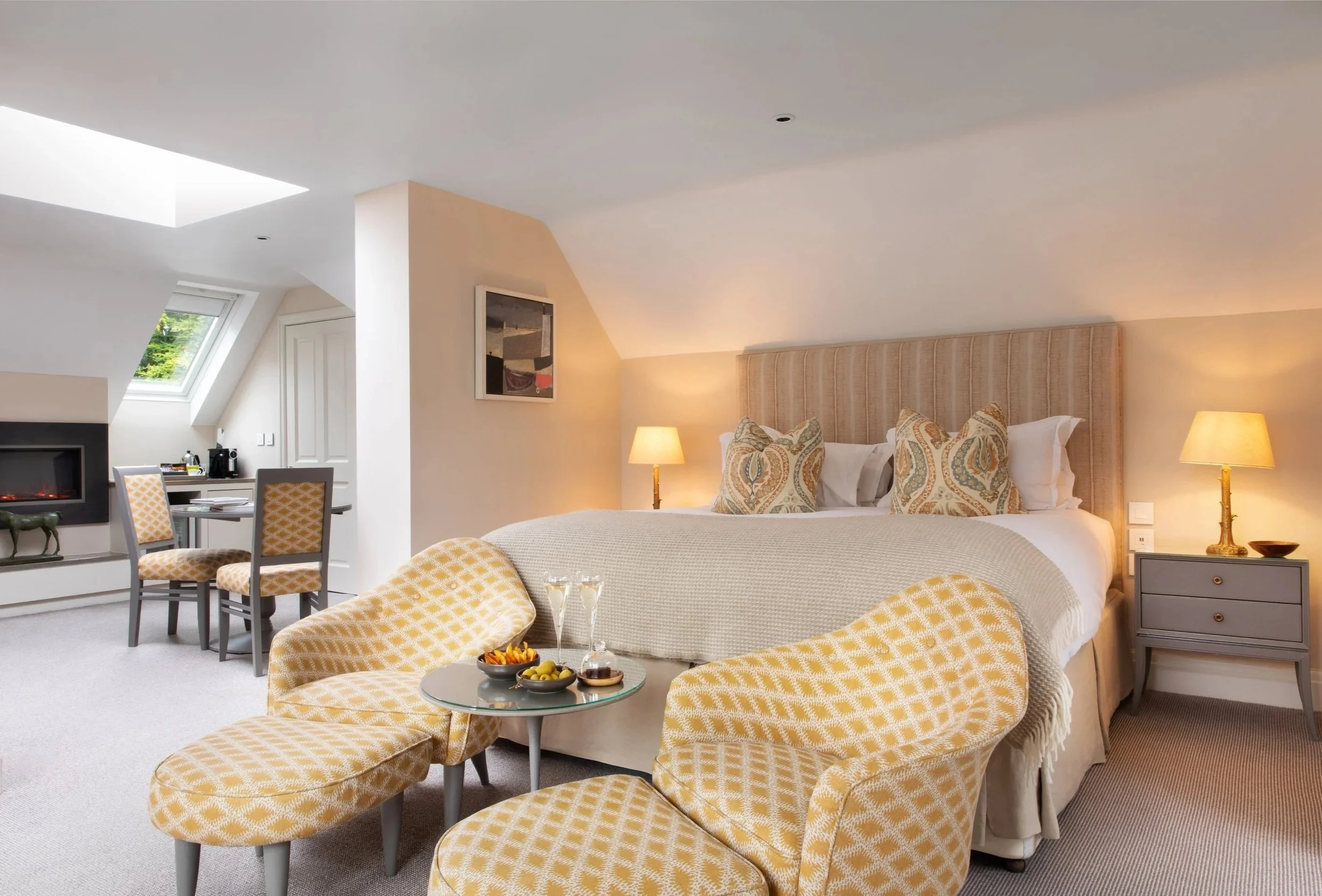 A cozy bedroom with a large bed, yellow patterned armchairs, and a small round glass table with snacks. The room has soft lighting from bedside lamps, a framed abstract artwork, a sloped ceiling, and windows allowing natural light.