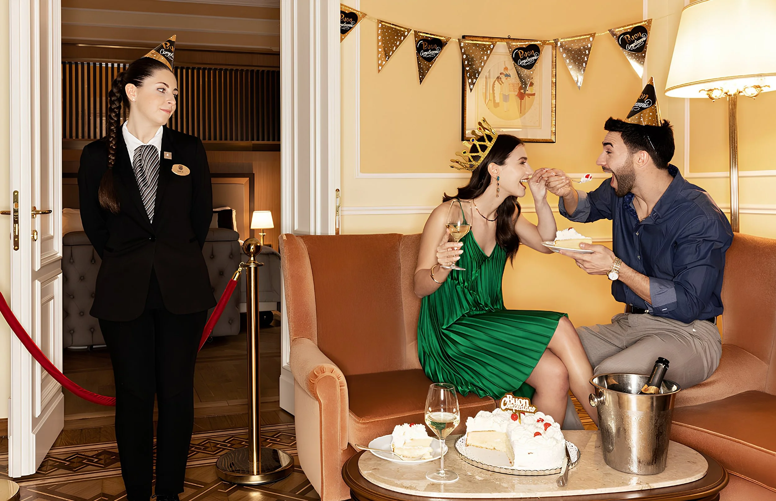 A woman in a green dress and a man with a cake slice and a wine glass celebrate a birthday, wearing a crown and party hat, with a waitress standing nearby at a hotel or restaurant party.