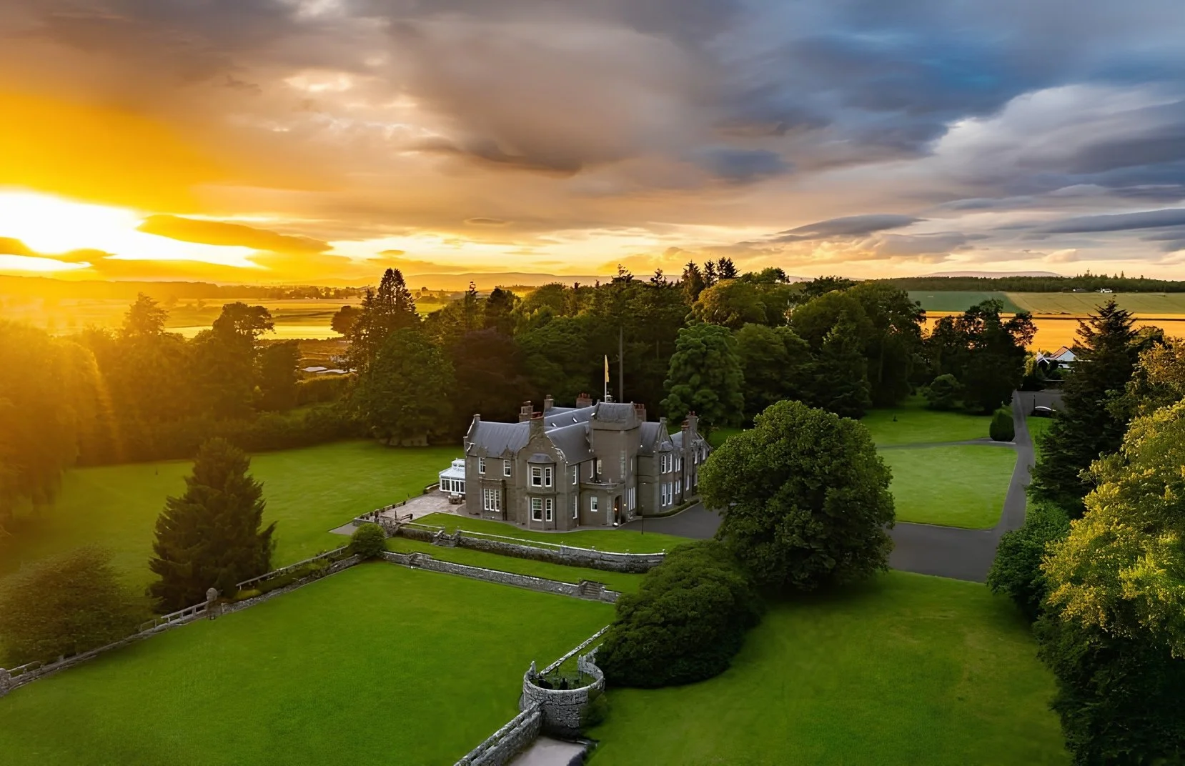 A large historic castle-style house with a turret, located on a well-manicured lawn surrounded by trees, with a sunset sky and rolling fields in the background.