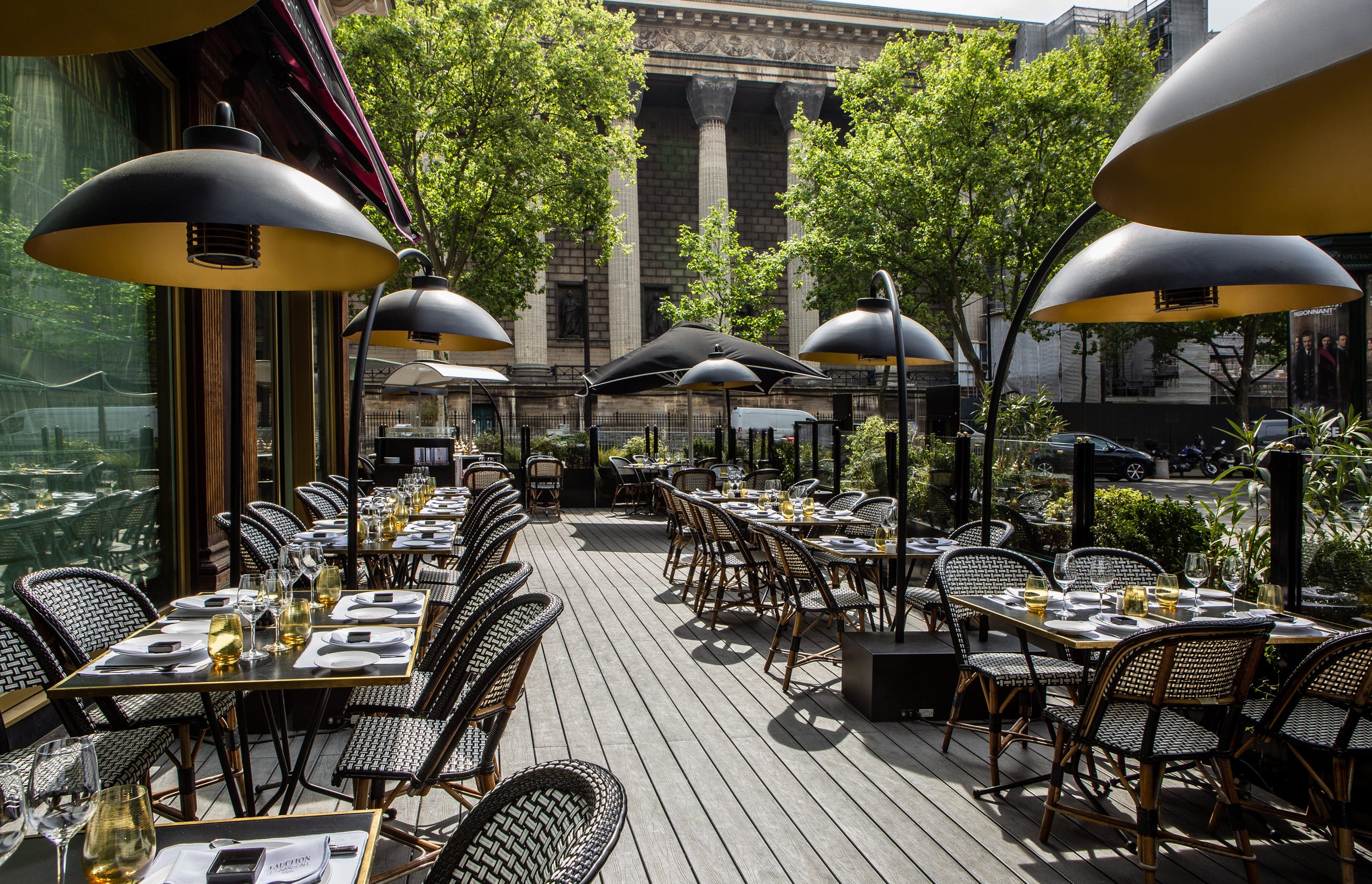 An outdoor restaurant patio with tables set for dining, including glasses and cutlery, under black and yellow umbrella shades, with trees and historic building in the background.